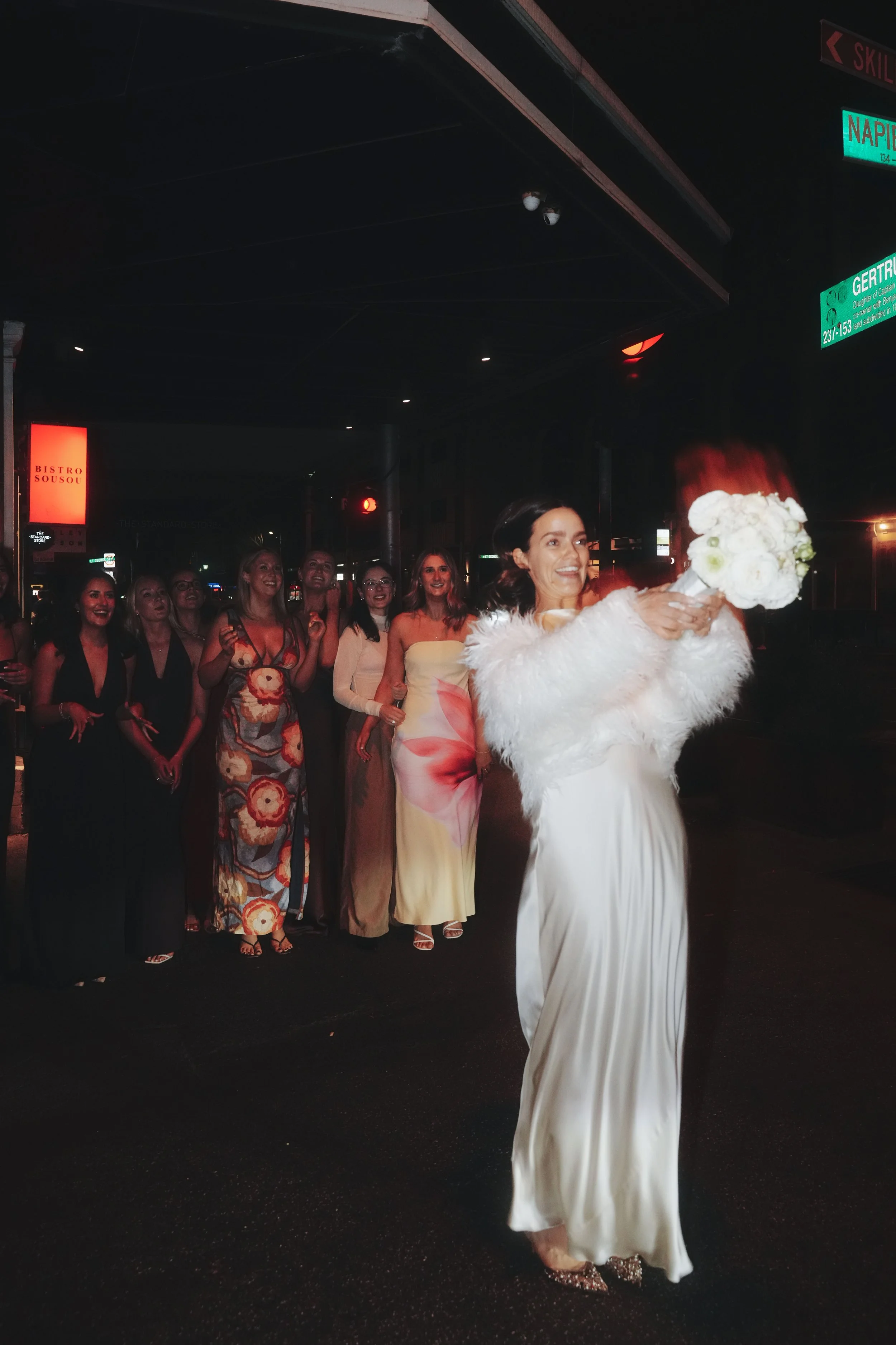 A woman in a white dress and fluffy shawl holding a bouquet of white flowers, smiling as she walks past a group of women in colorful dresses on a city street at night.