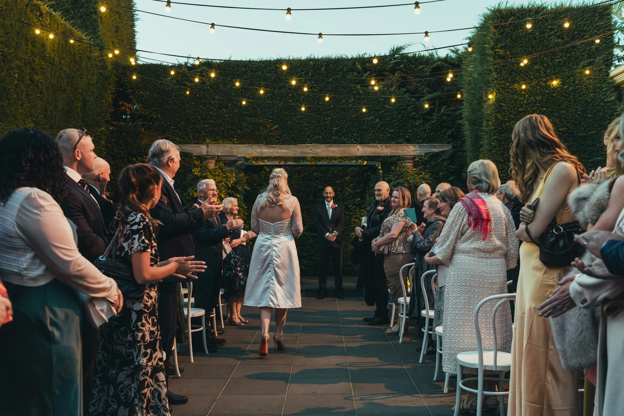 People attending an outdoor wedding ceremony at dusk, with string lights overhead, surrounded by tall green hedges.