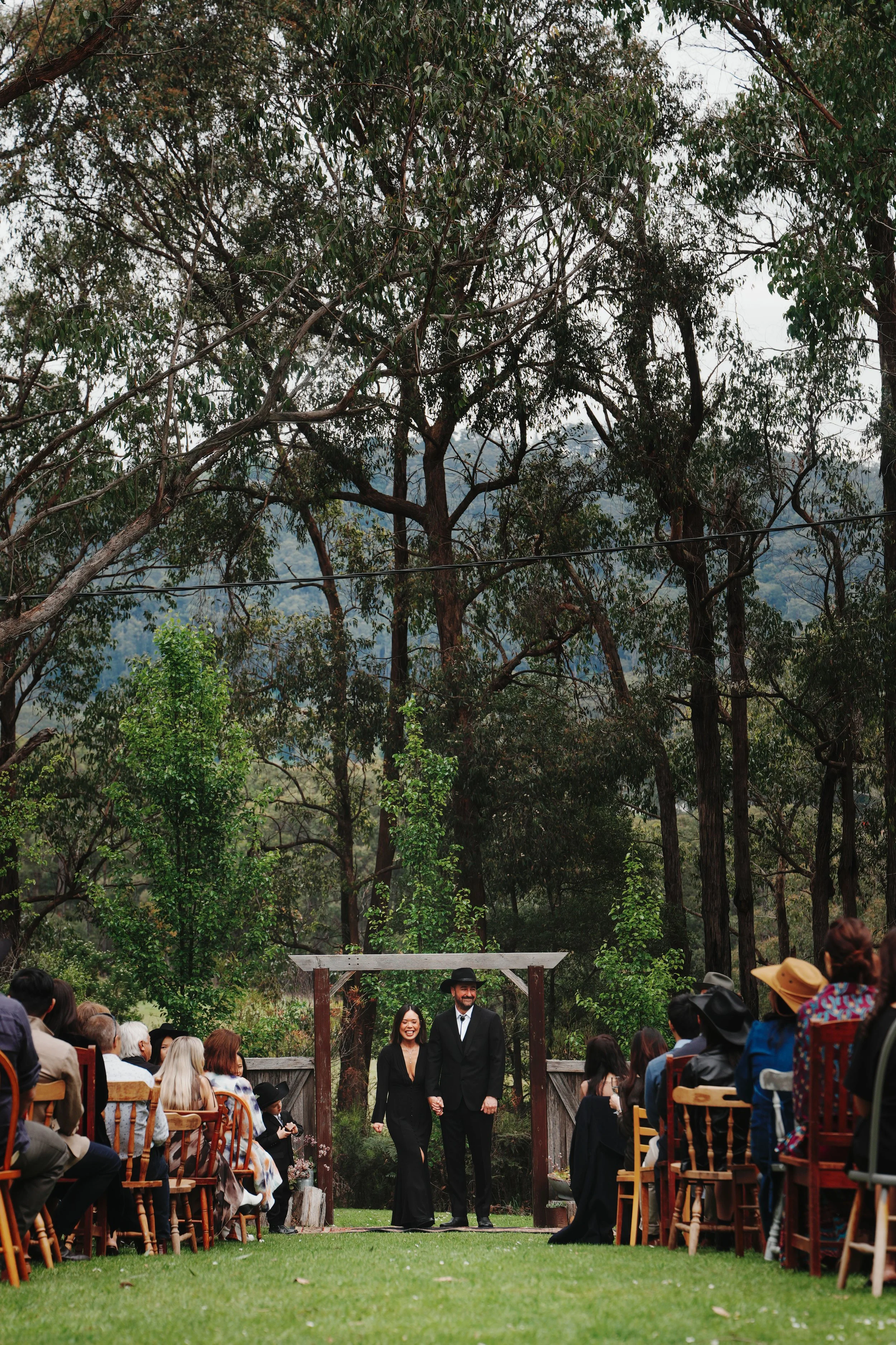 A wedding ceremony outdoors in a forest with guests sitting on wooden chairs, a couple holding hands walking down the aisle, surrounded by trees and mountains in the background.