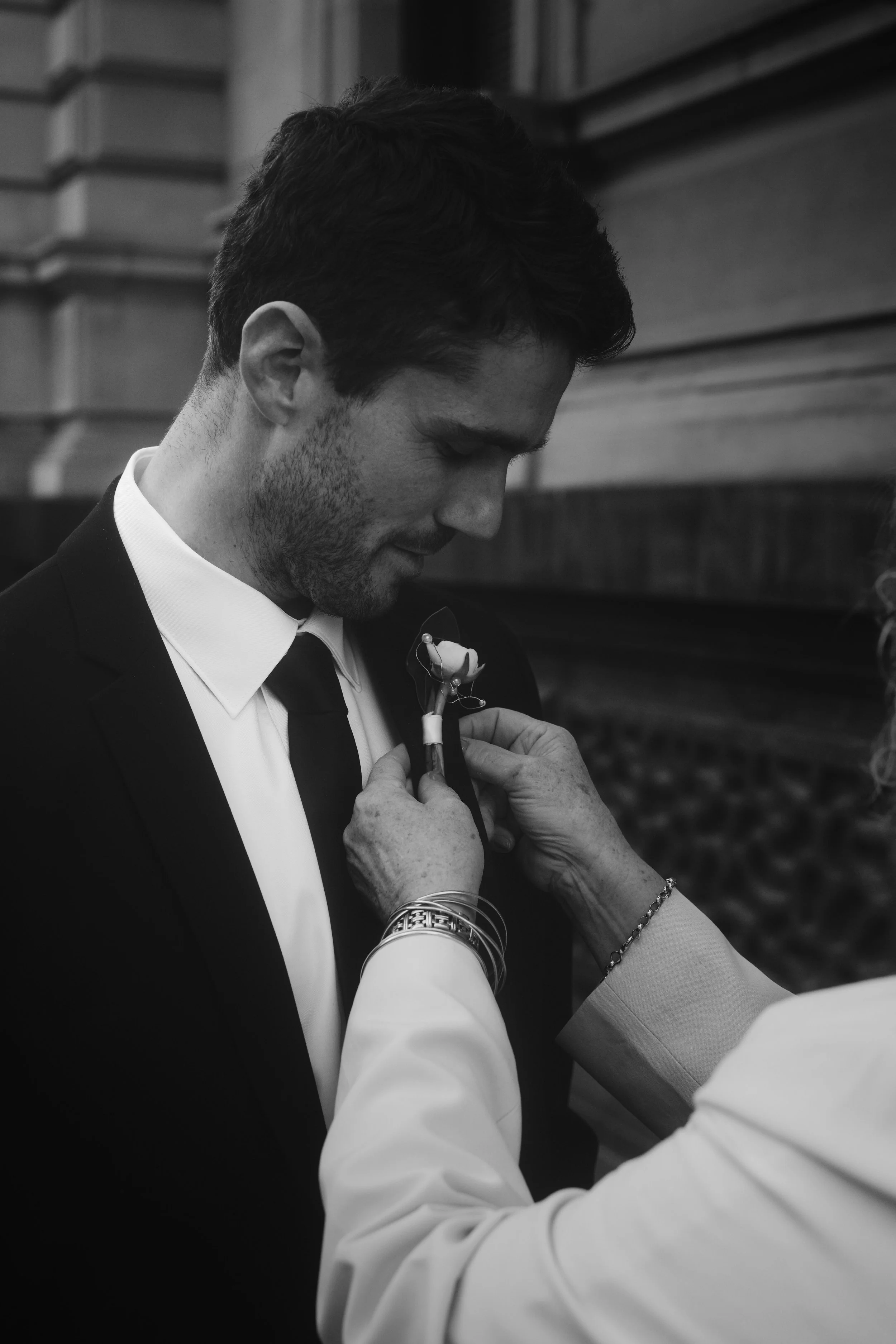 A man in formal wear, possibly at a wedding, is being helped to put on a boutonniere or flower pin on his suit lapel by a woman.