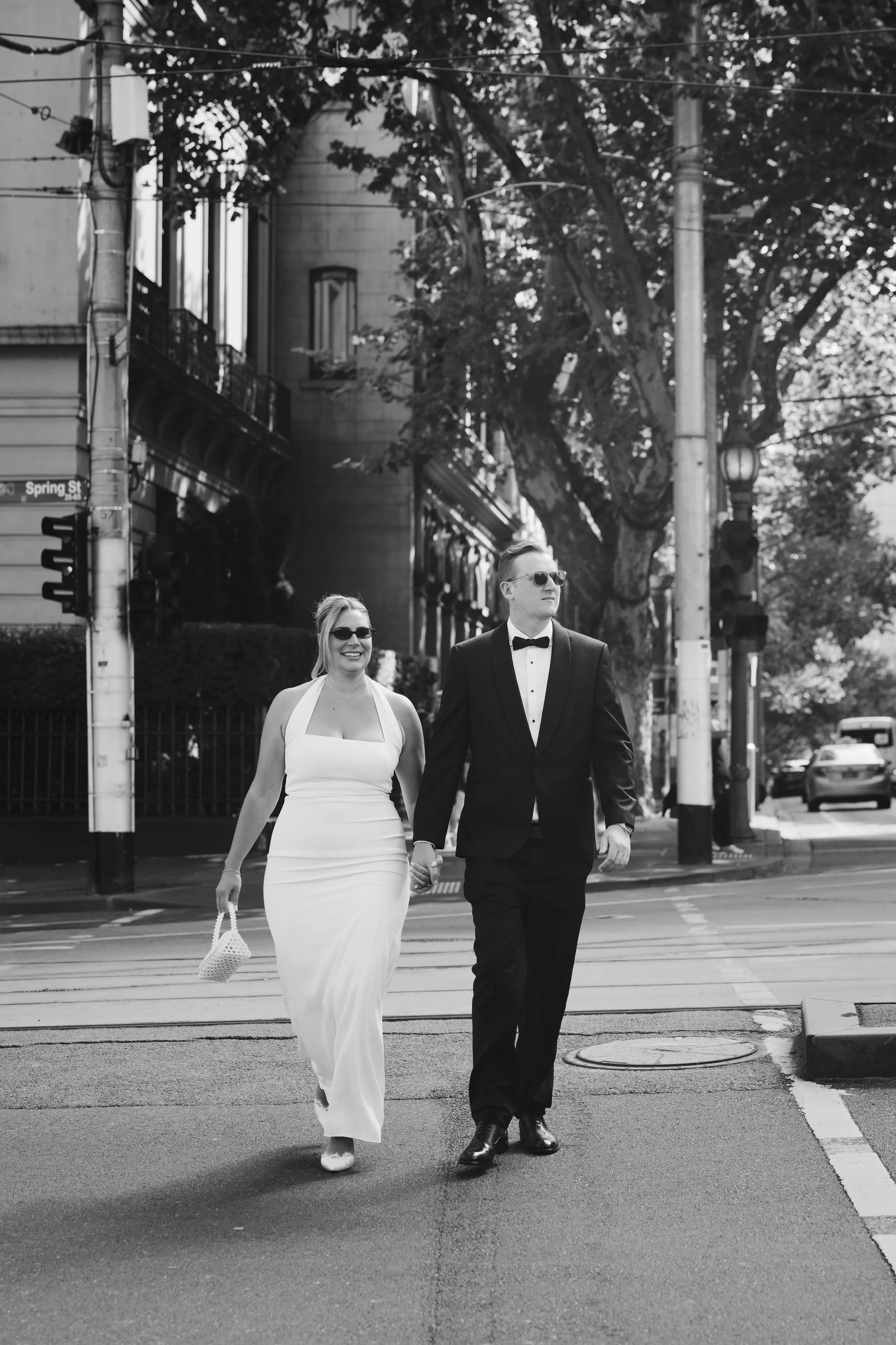 A couple dressed in formal attire walking hand in hand across a city street. The woman is in a white gown and sunglasses, carrying a small purse. The man is wearing a tuxedo and sunglasses. There are trees, streetlights, and buildings in the backgrou