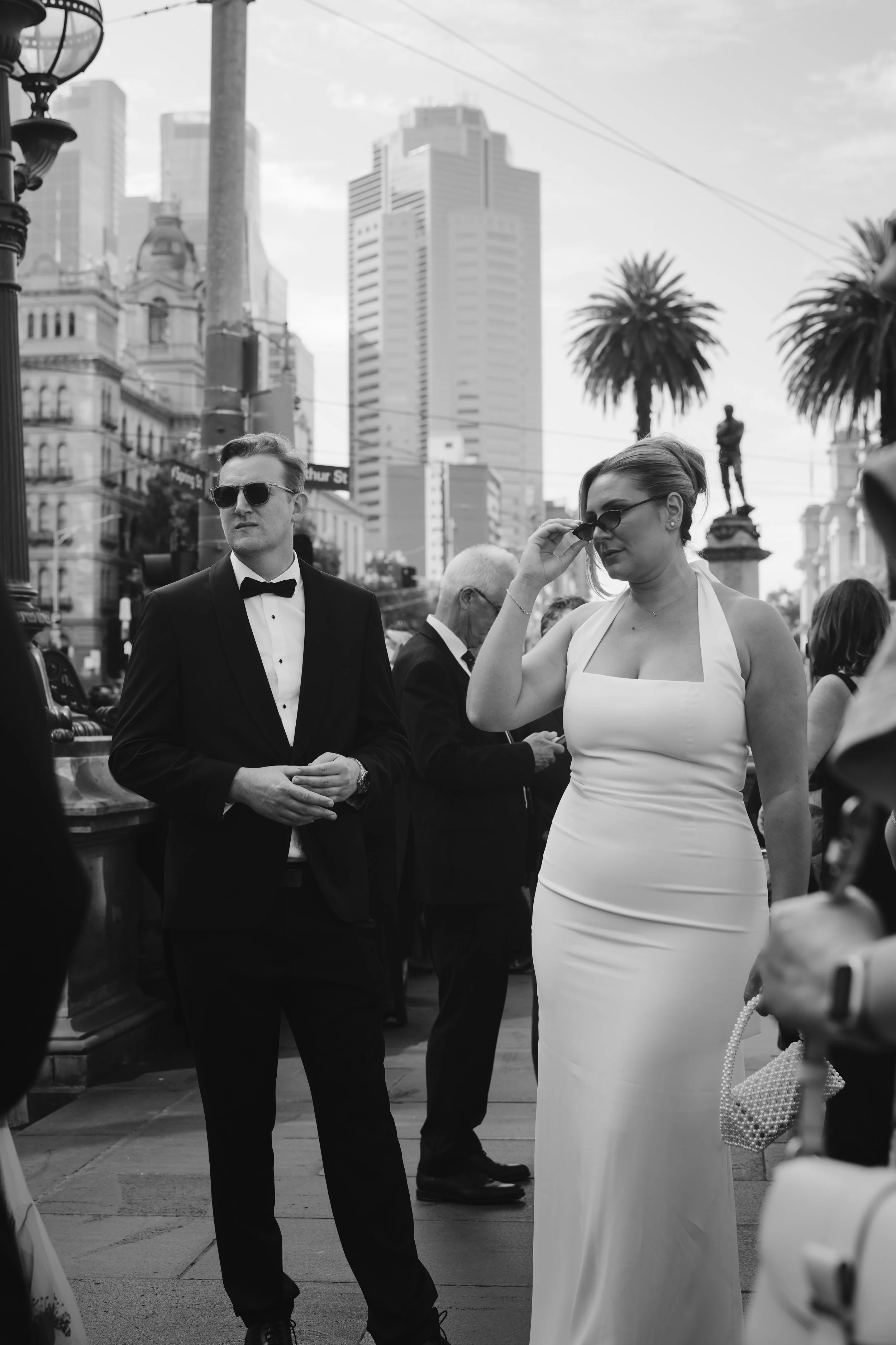 A black and white photo of a man in a tuxedo and sunglasses and a woman in a white dress with glasses, standing outdoors in an urban setting with skyscrapers and palm trees in the background.