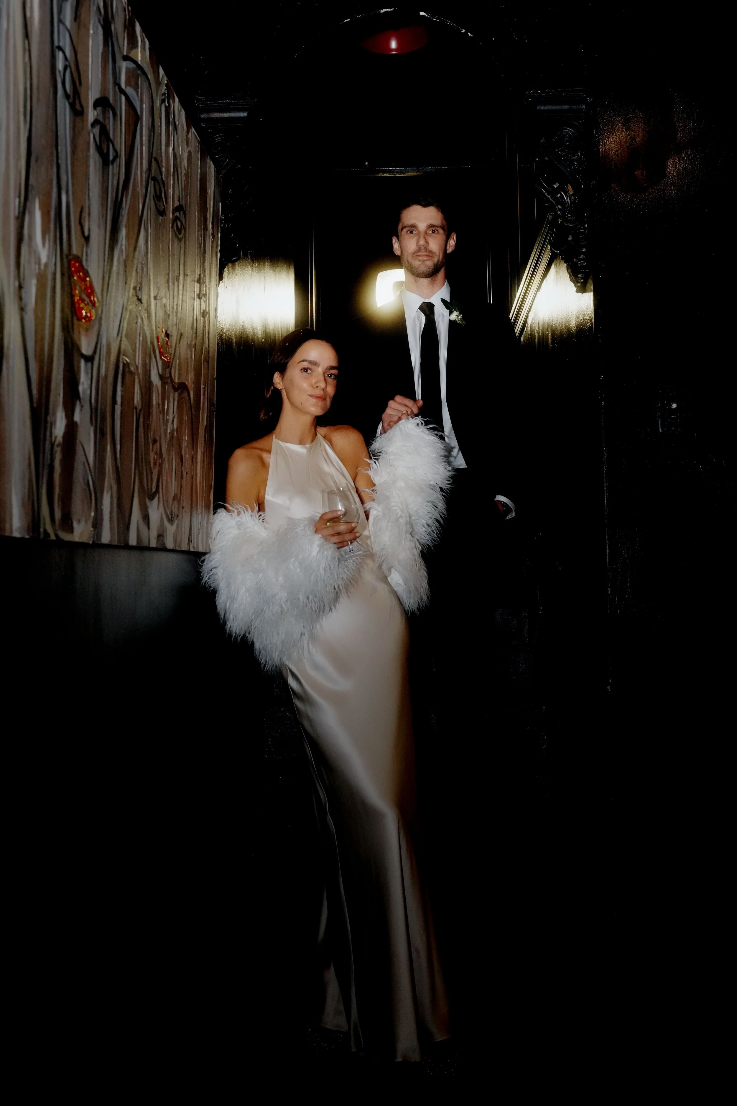 A man and woman in formal attire at a wedding reception, the woman holding a glass of wine, standing against a dark wall with ornate woodwork and dim lighting.
