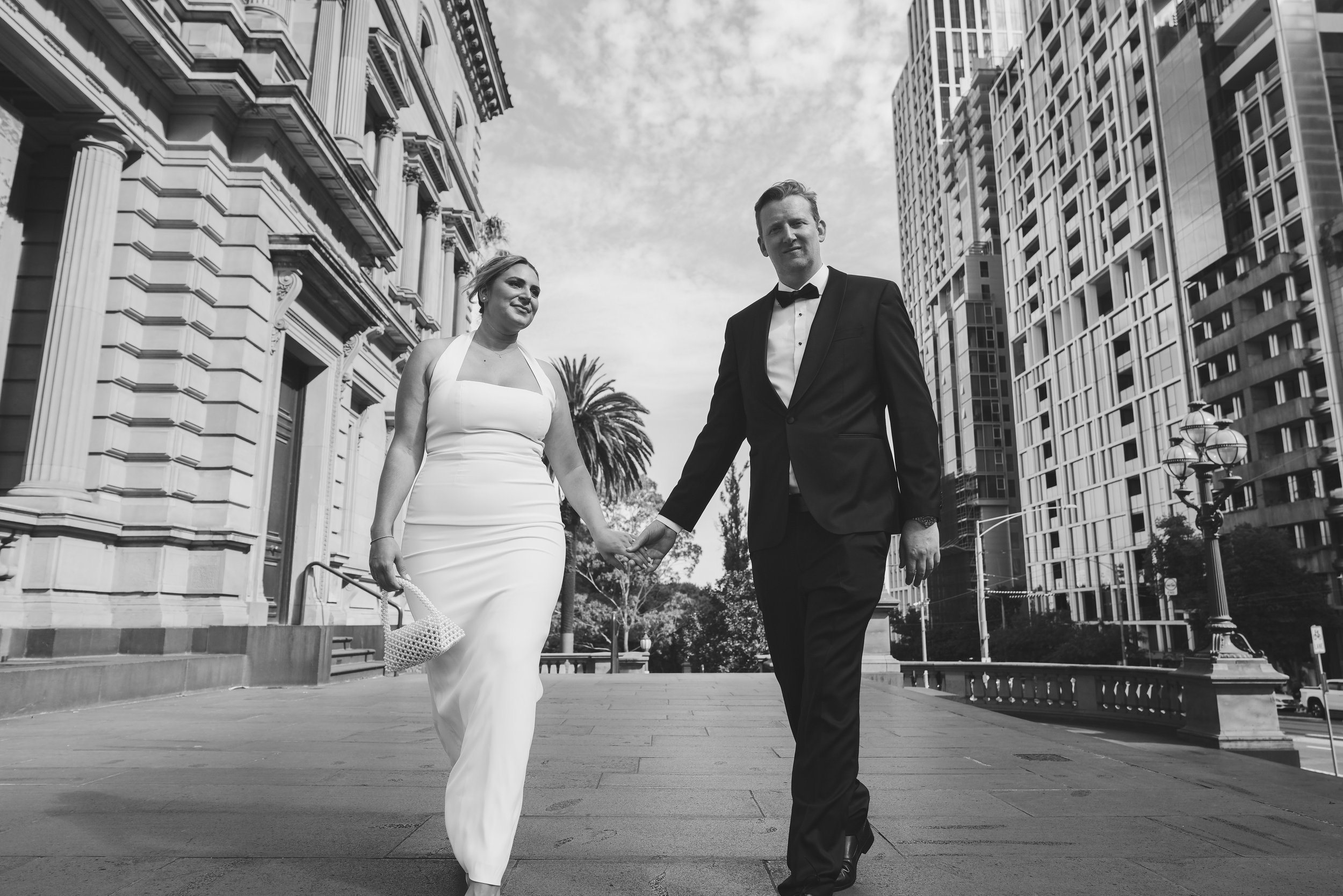 A bride and groom walking hand in hand in an urban area with tall modern buildings and a historic building with columns. The bride wears a white gown, and the groom is dressed in a black tuxedo. The sky is partly cloudy.
