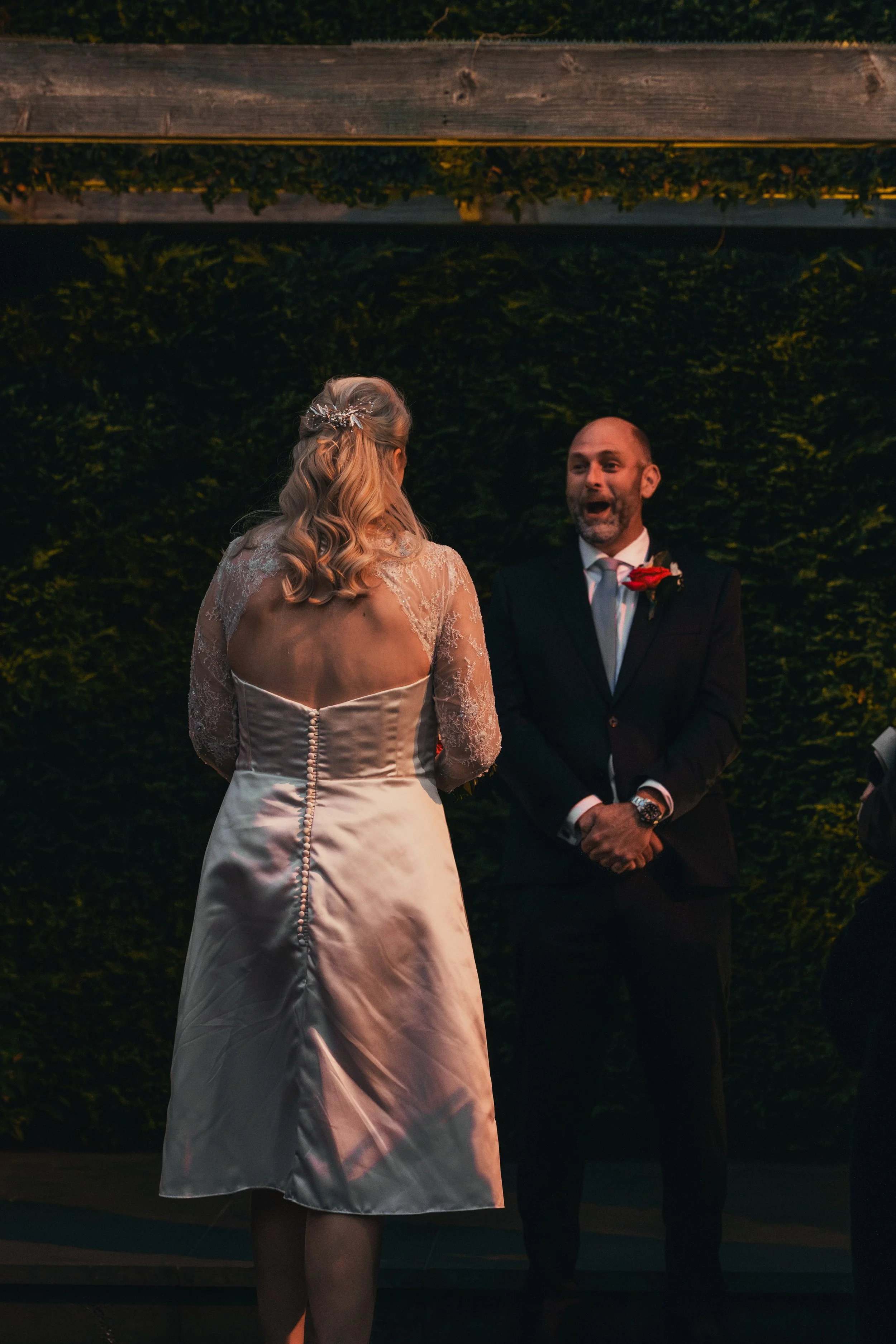 A bride and groom exchanging vows at an outdoor wedding ceremony during the evening. The bride is facing away from the camera, wearing a white satin wedding dress with lace details, while the groom is facing her, dressed in a dark suit with a white s