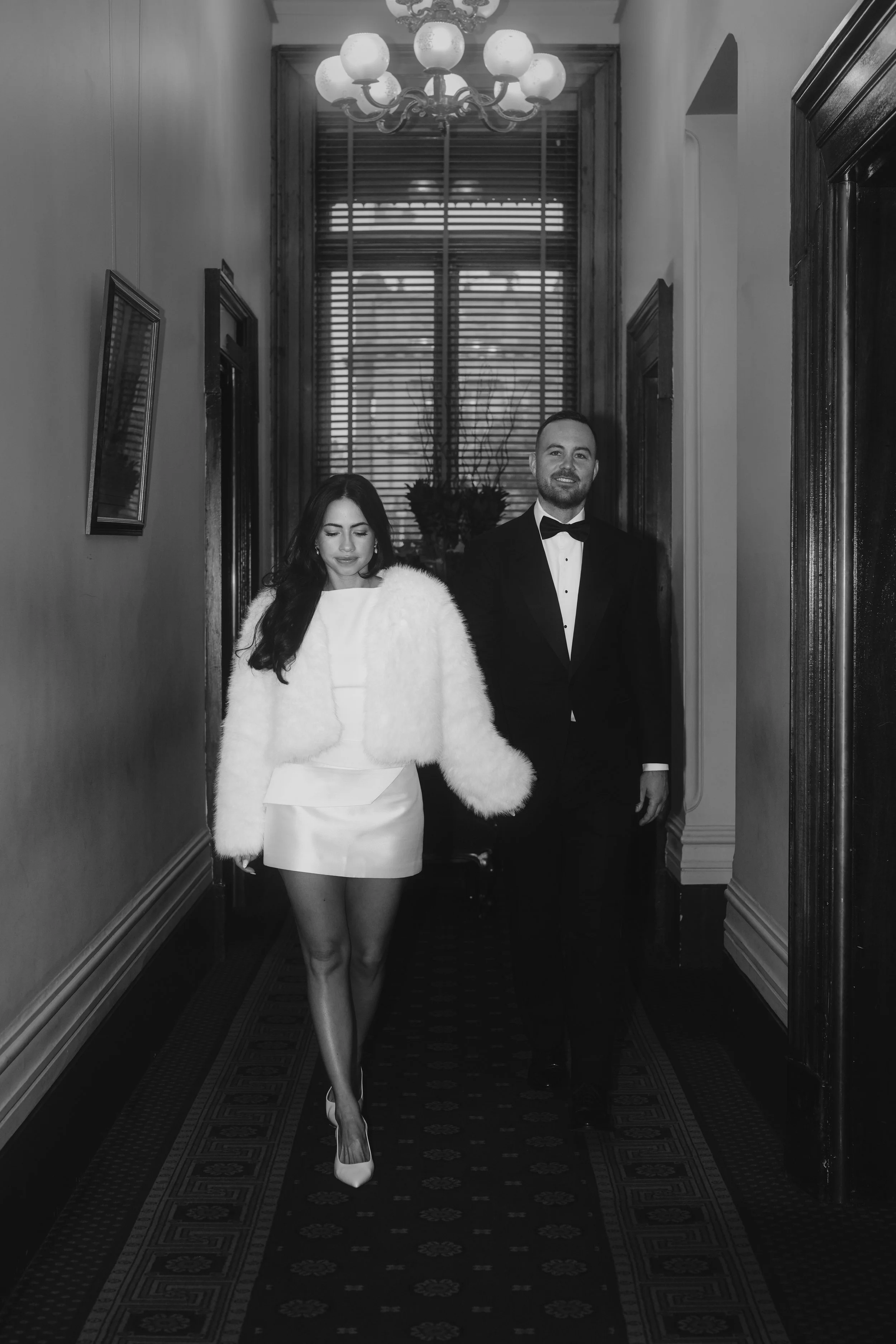 A woman in a white dress and high heels walking down a patterned corridor with a woman in a tuxedo following behind her. The woman in white has long dark hair, wearing earrings and a fluffy white coat. The man has a beard and mustache, dressed in a t