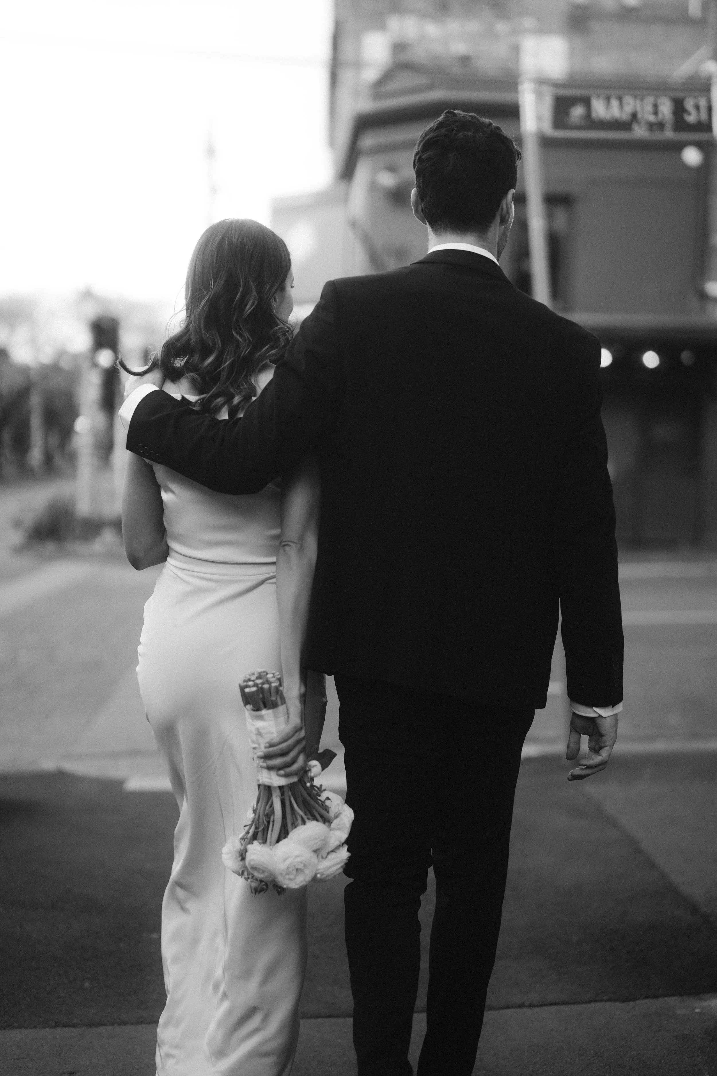 A black and white photo of a man and woman walking arm in arm in an urban setting, with the woman holding flowers and a stuffed animal.