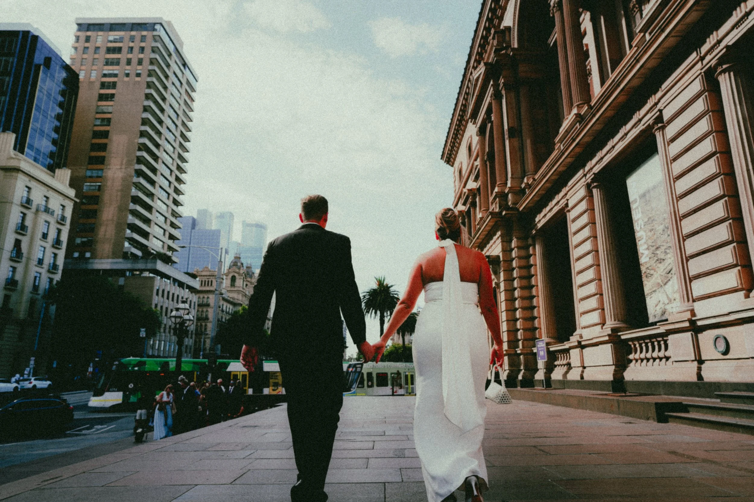 A bride and groom holding hands and walking in an urban city street, with tall buildings and a city bus visible in the background.