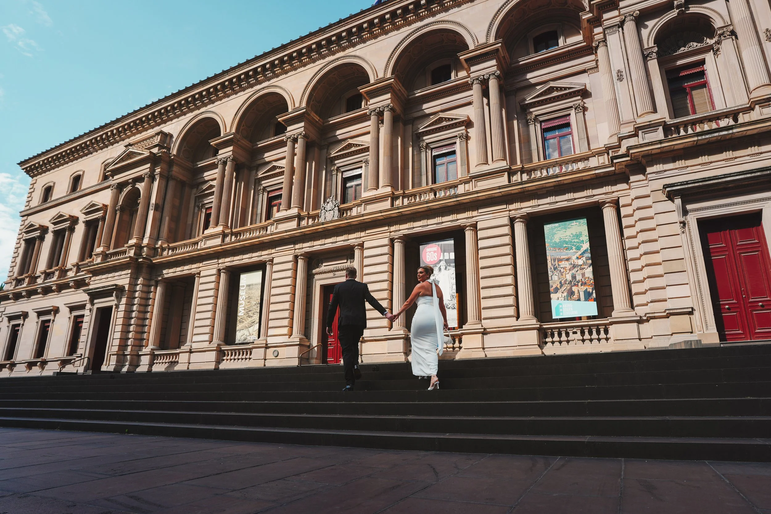 A couple dressed in formal wedding attire holding hands and walking up the steps outside a grand historic building with ornate architectural details and large arched windows.