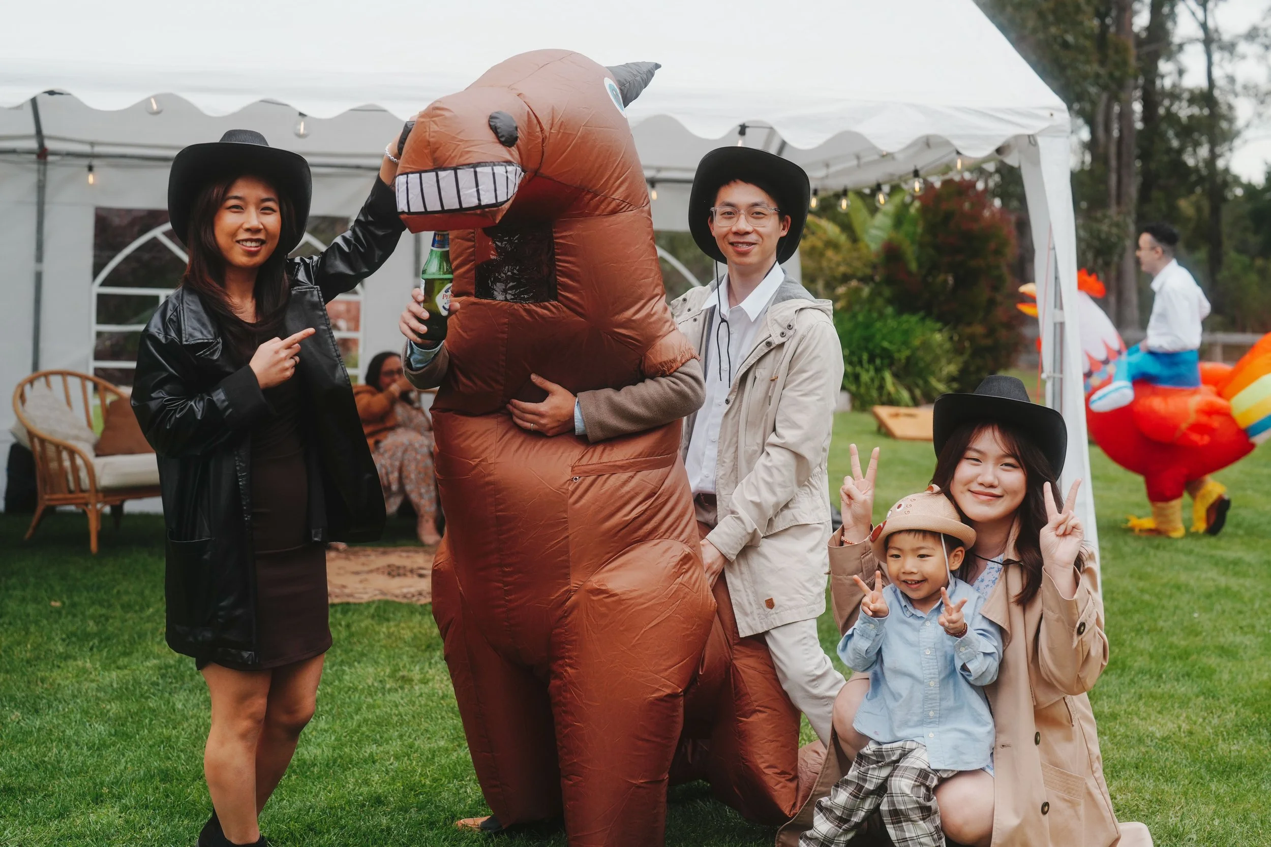 Group of people at outdoor event with Halloween costumes, including a person in a dinosaur costume, others in cowboy hats, and children making peace signs.