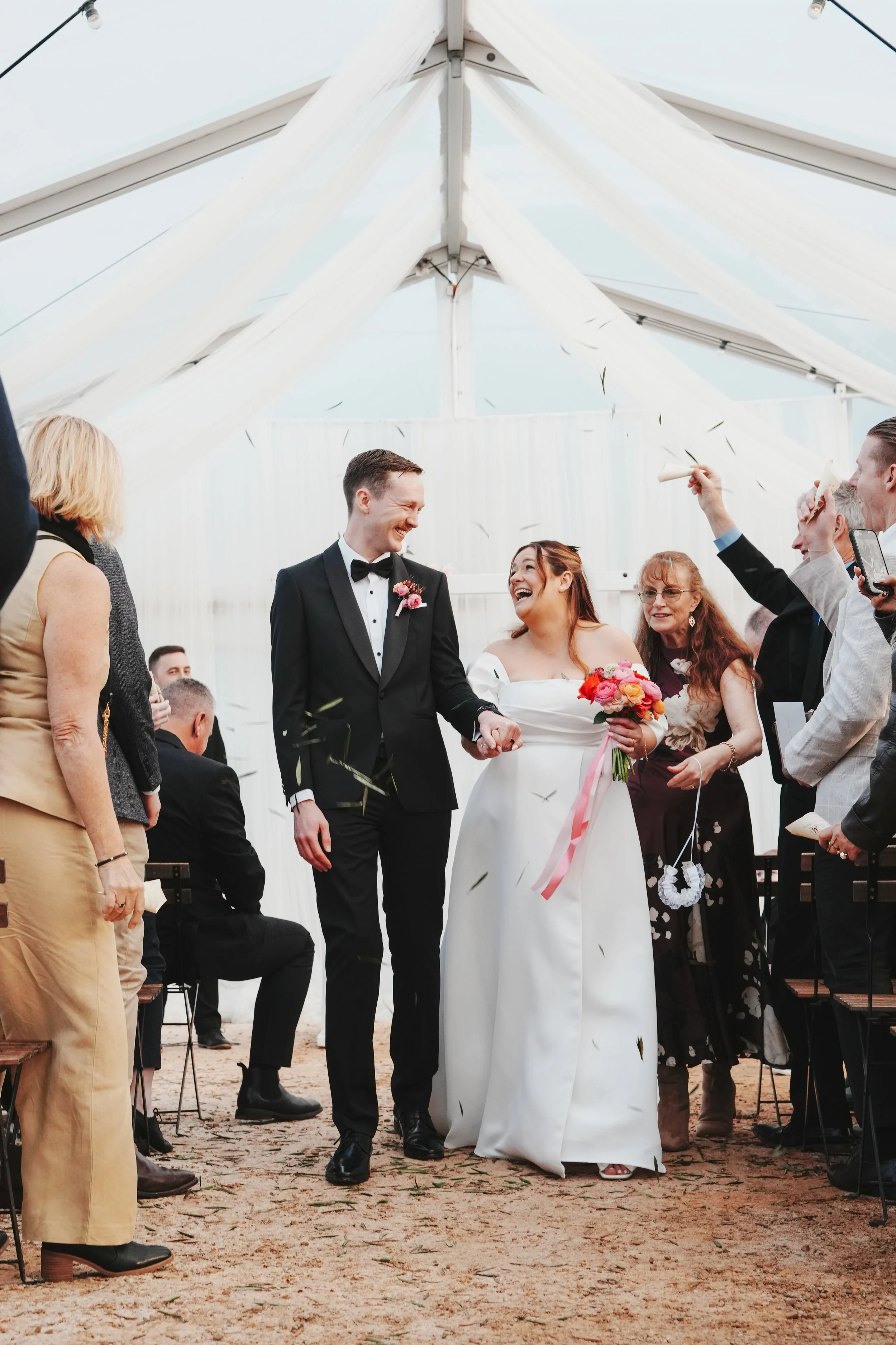 A wedding ceremony inside a white tent. A bride in a white dress holding a bouquet of pink and red flowers, and a groom in a black tuxedo are holding hands and smiling at each other. Guests are seated and standing around, some tossing confetti, celeb