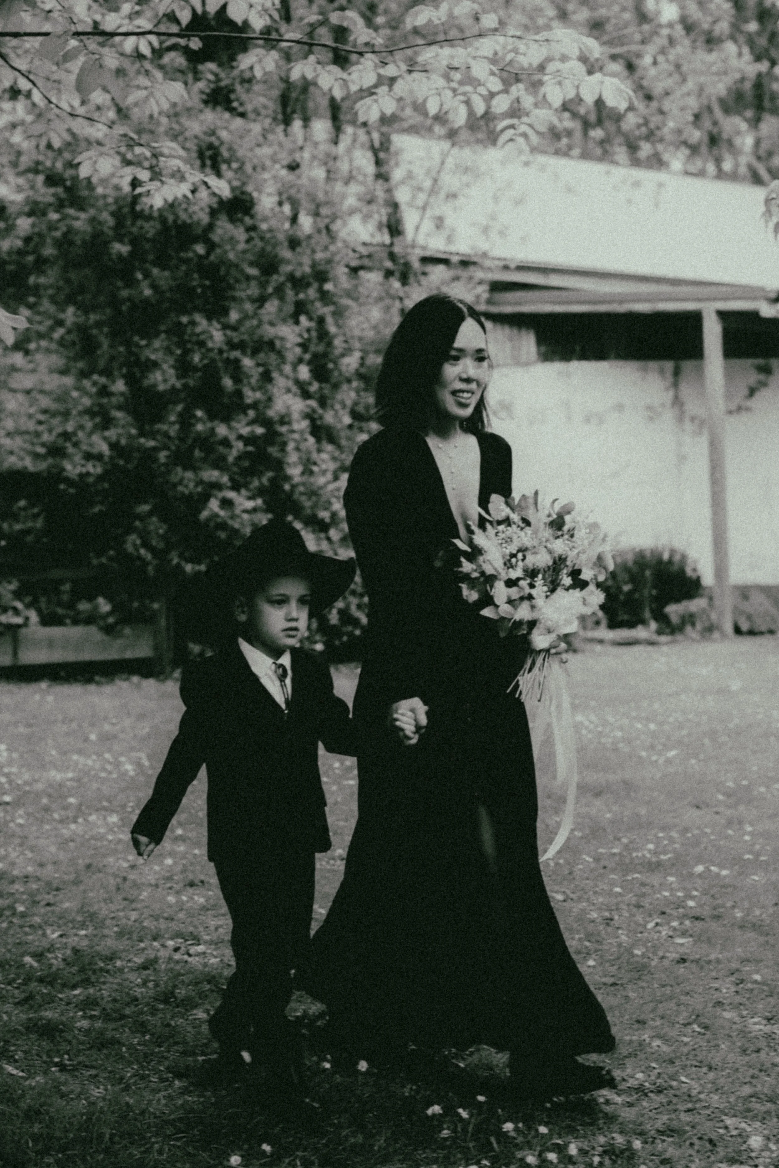 A woman wearing a black dress holding a bouquet of flowers walking with a young girl dressed in black and a large hat in an outdoor rural setting, with a white building and trees in the background.