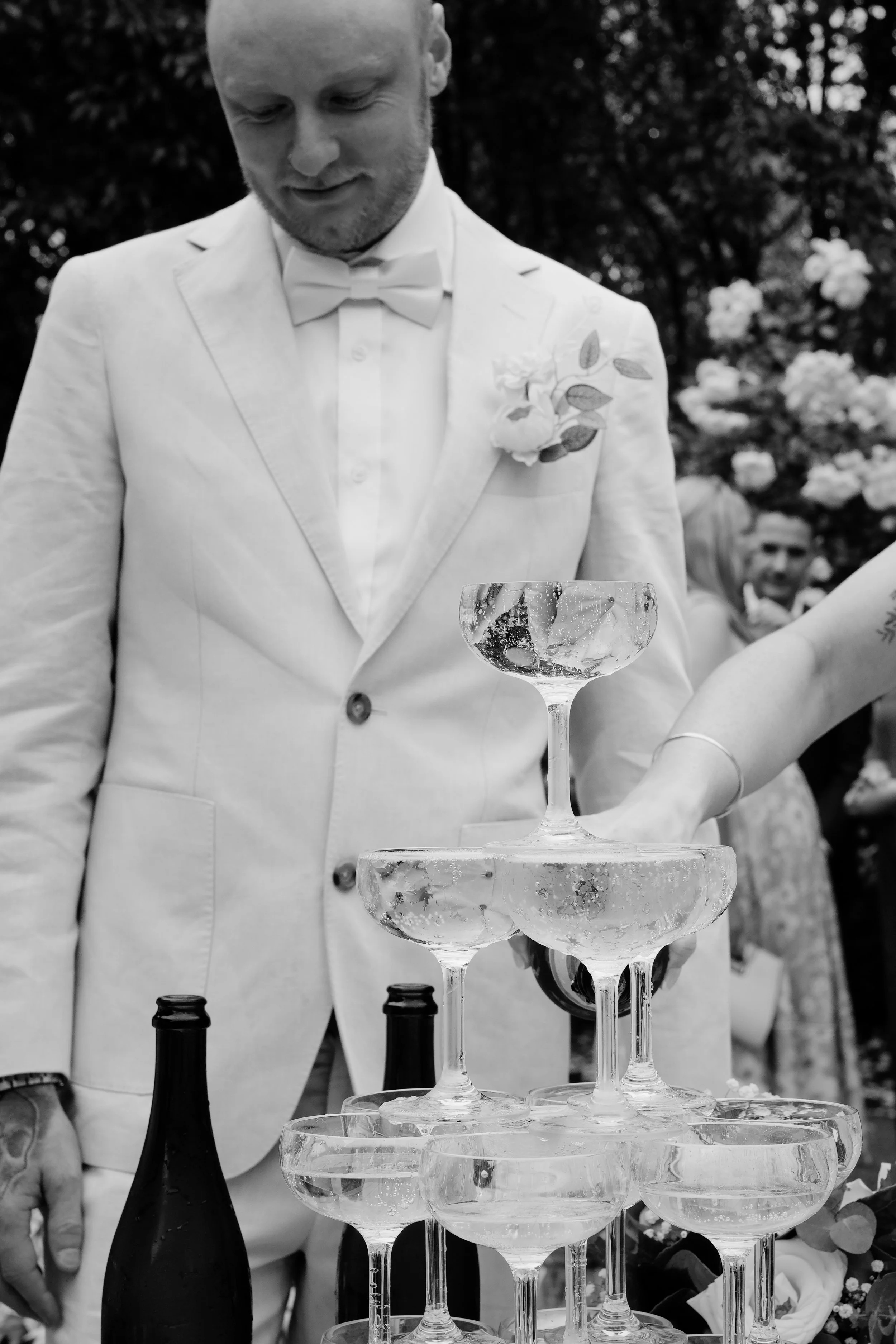 A groom dressed in a white tuxedo with a bow tie at a wedding reception, focusing on a champagne tower.