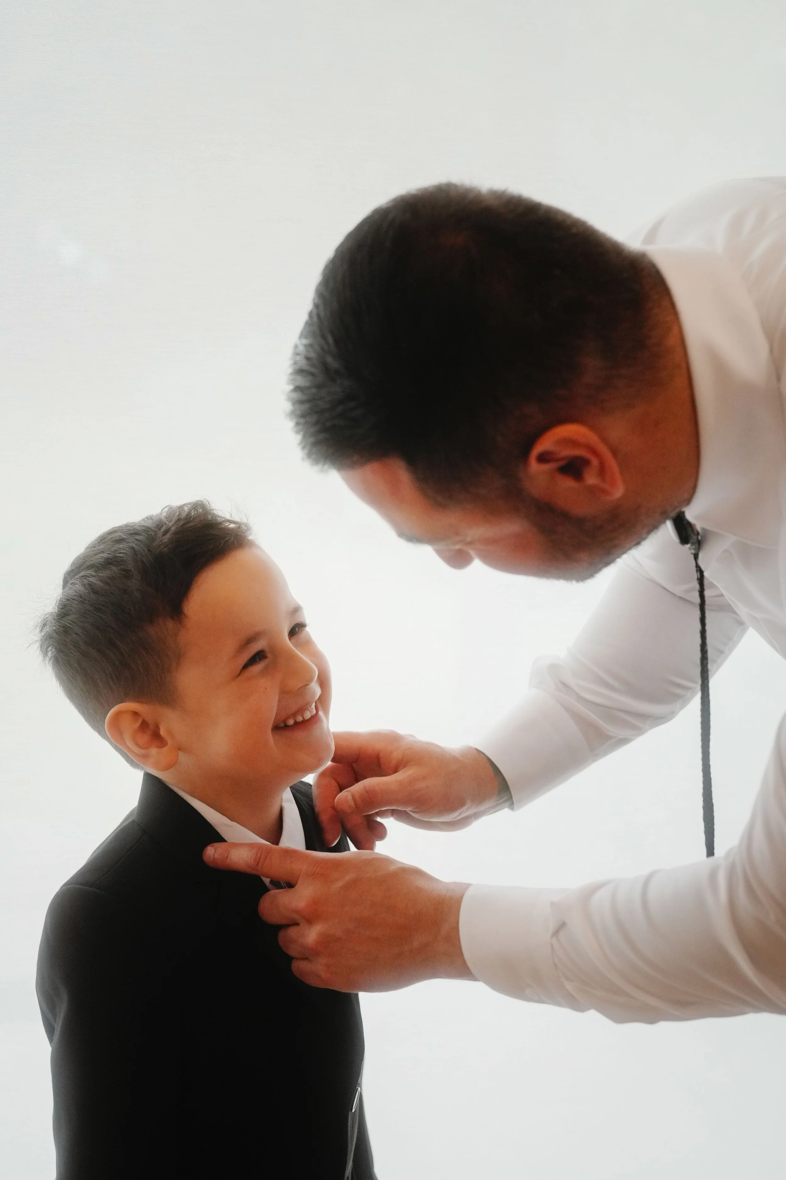 A man in a white shirt and a young boy in a black suit are smiling at each other. The man is bending down slightly, touching the boy's shoulder, and they are looking at each other in a moment of connection and warmth. The background is white and plai