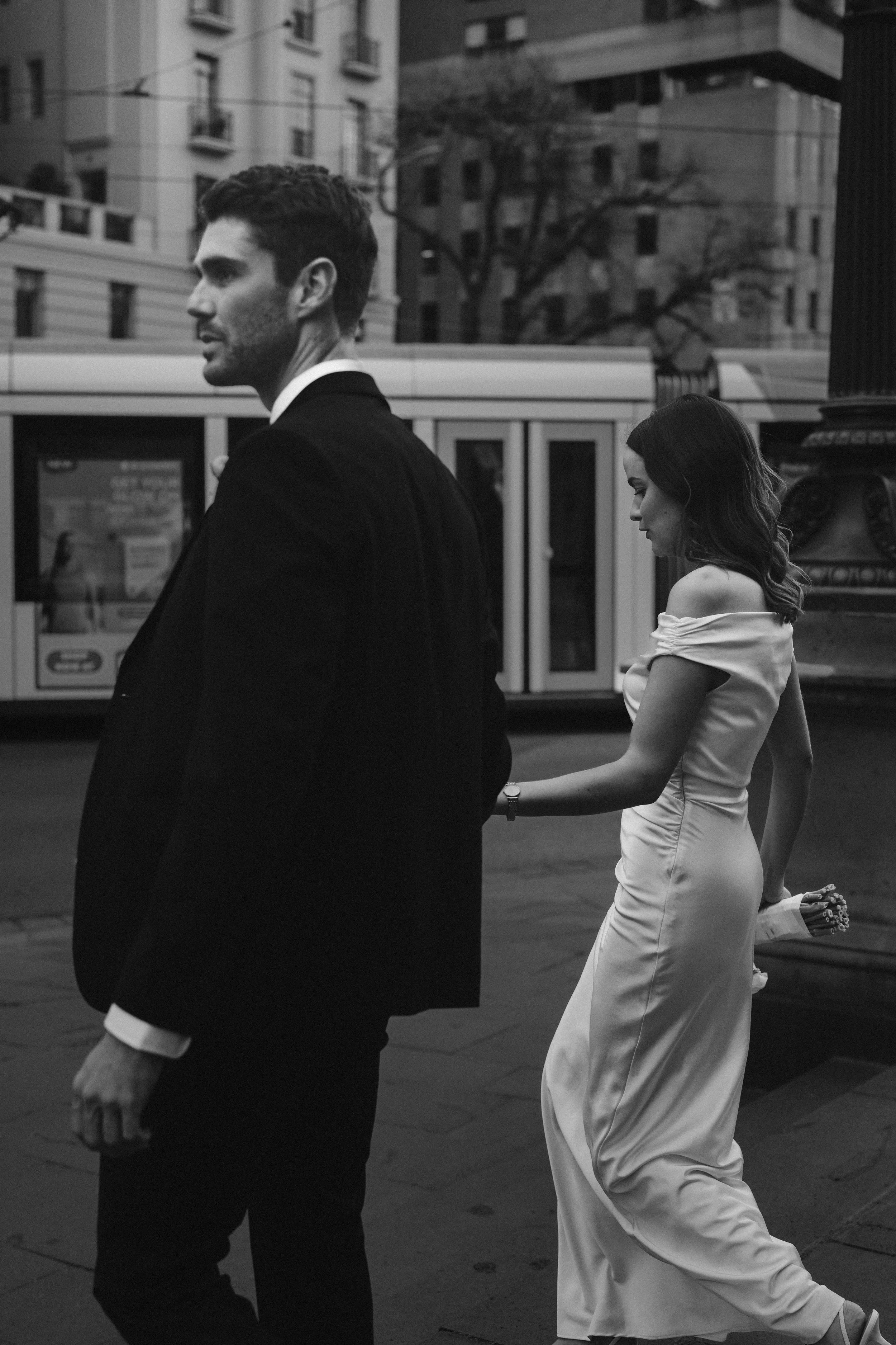 A man in a tuxedo and a woman in a satin dress holding hands and walking outdoors in an urban street with buildings and trees in the background.