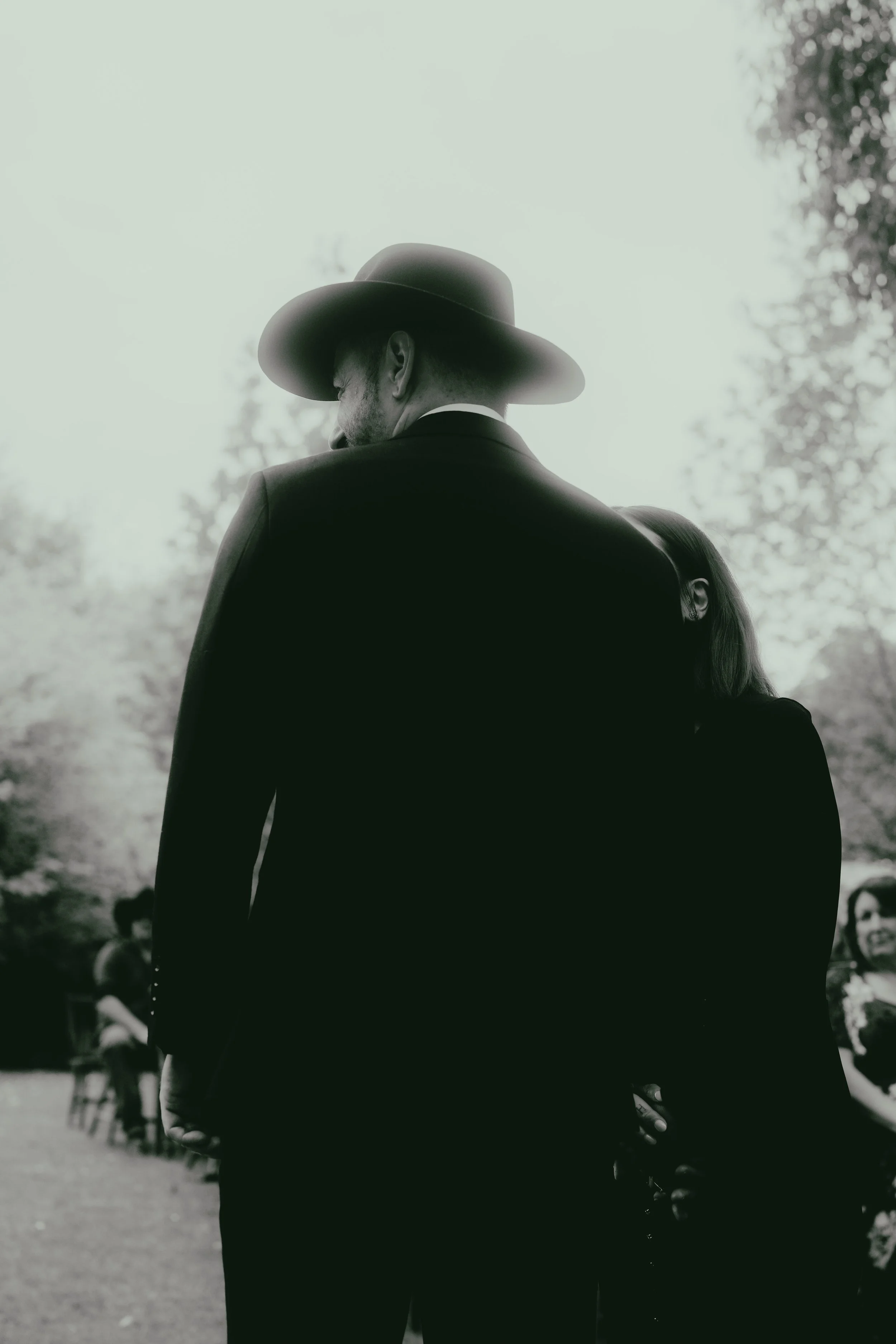 Silhouette of a man wearing a wide-brimmed hat, viewed from behind, outdoors with blurred trees and people in the background.