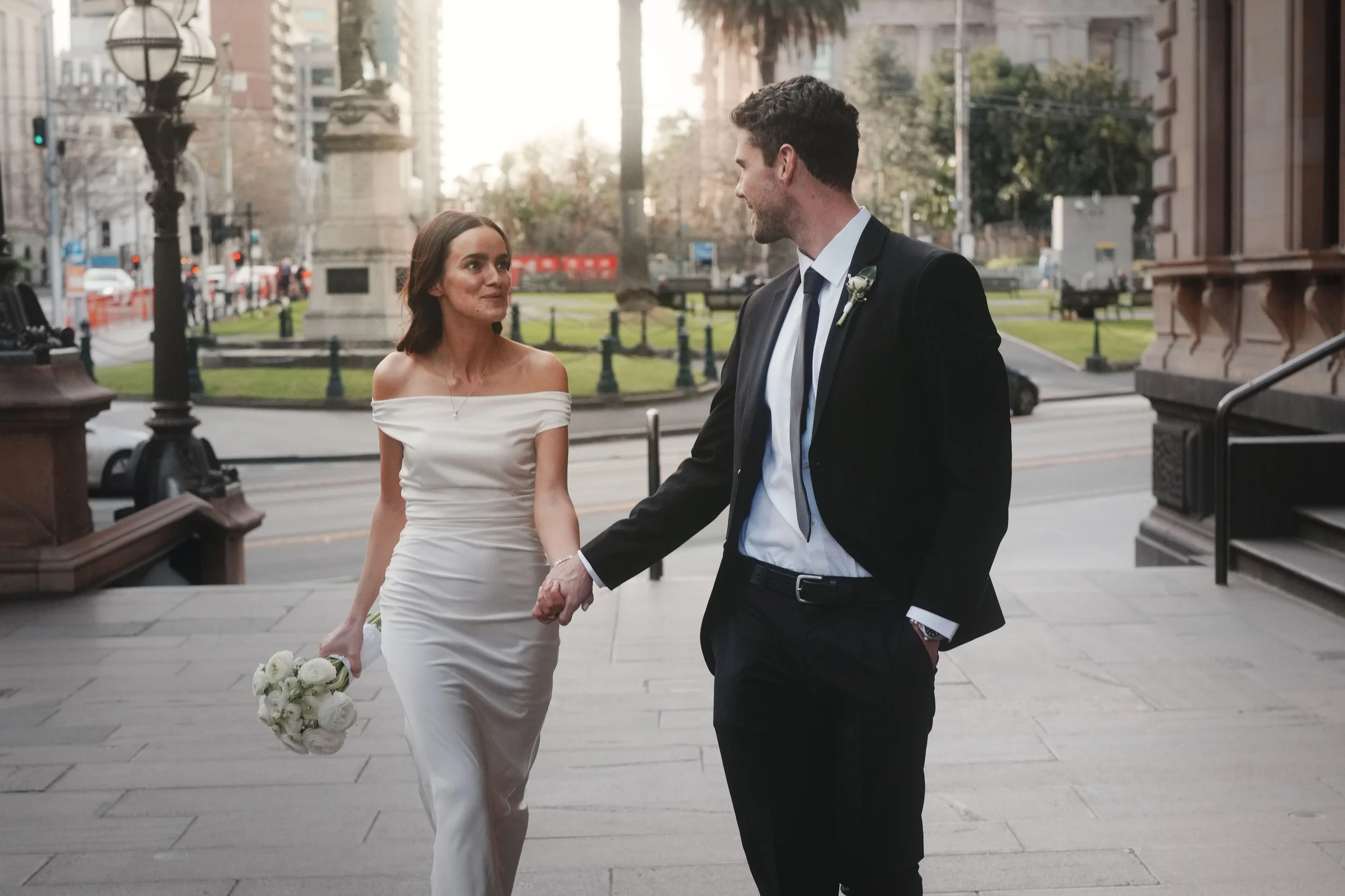 A newlywed couple walking hand in hand outdoors in a city, with the woman holding a bouquet of white roses and the man wearing a black suit with a boutonniere, smiling at each other.