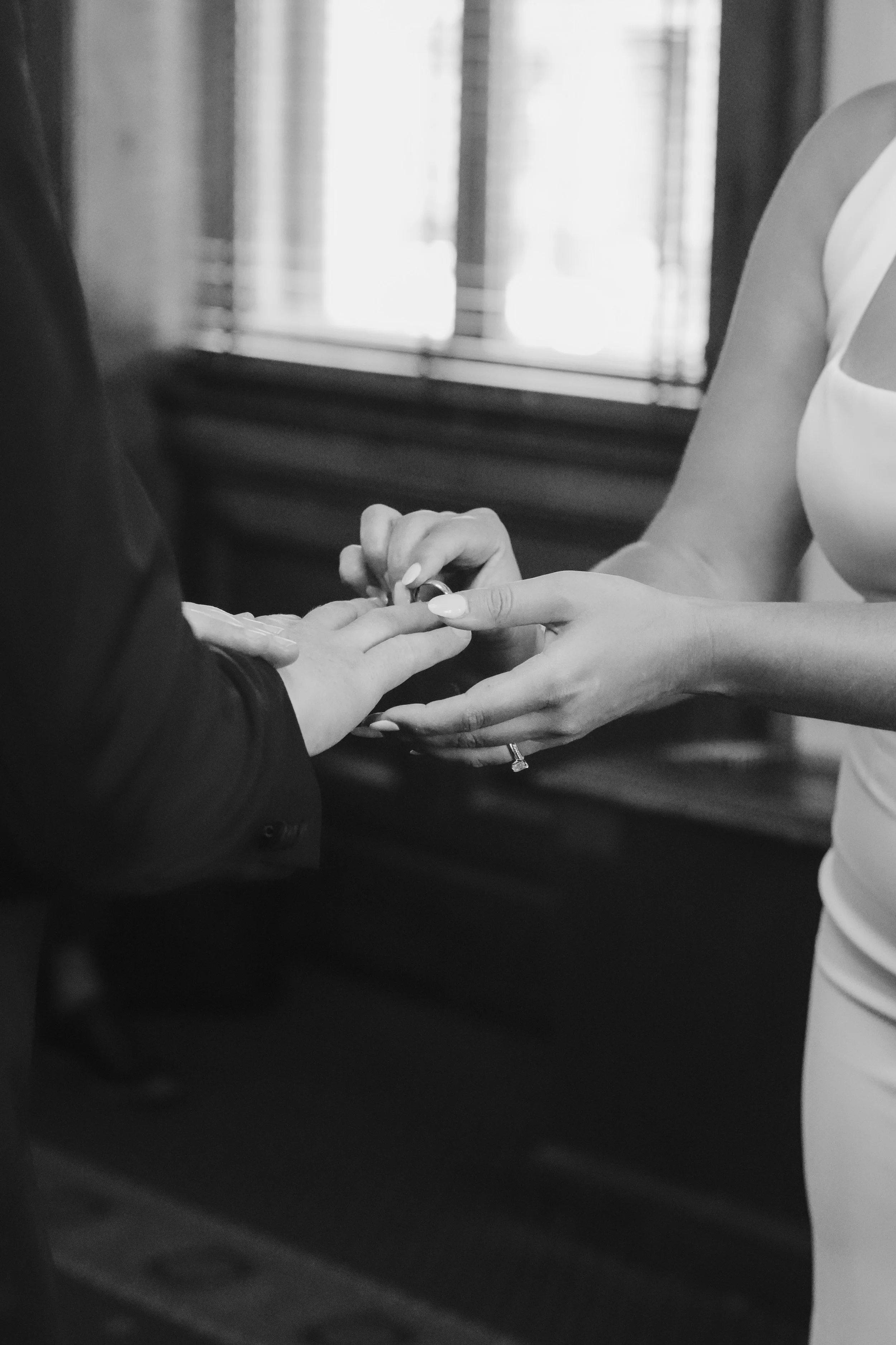 A person in a dark suit is placing a ring on another person's finger, whose hand is extended. Another person, possibly a woman, is supporting the hand with the ring, wearing a ring on her left ring finger. The background shows a window with curtains.