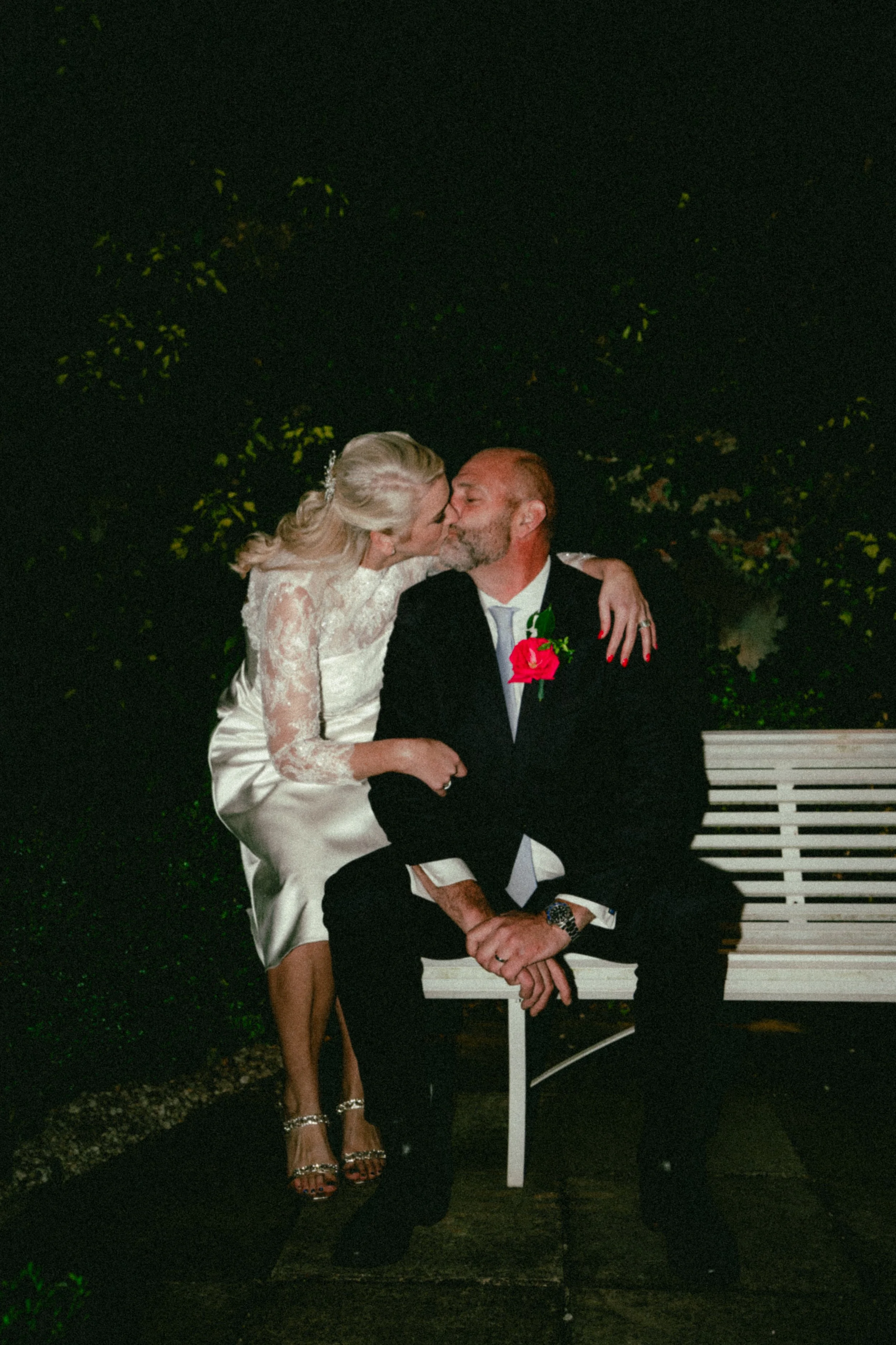 A woman in a white lace dress and high heels kisses a man in a suit with a red rose boutonniere on a park bench at night.