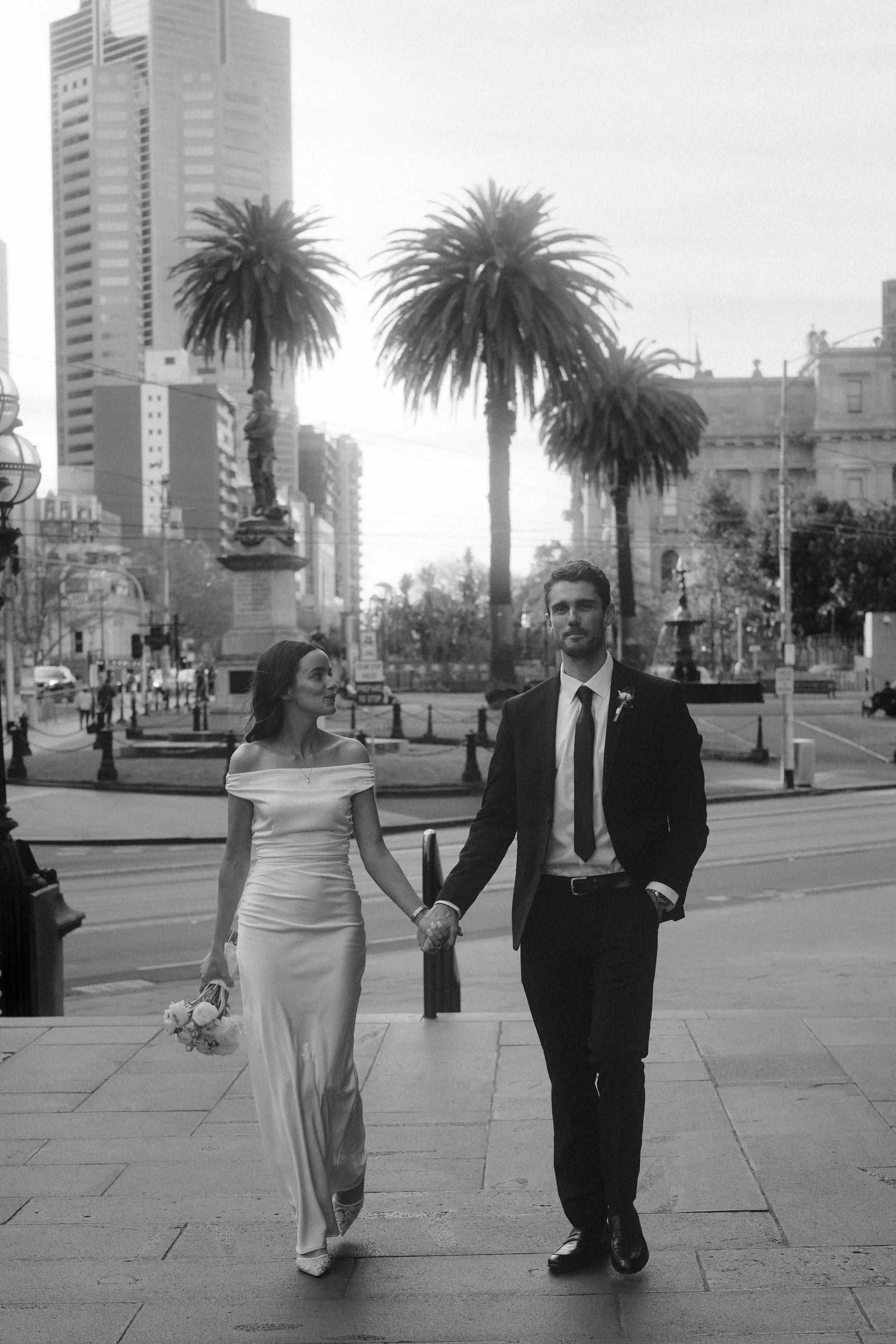 A man and woman dressed in wedding attire holding hands and walking outdoors in a city with tall buildings, palm trees, and streetlights, with other people in the background.