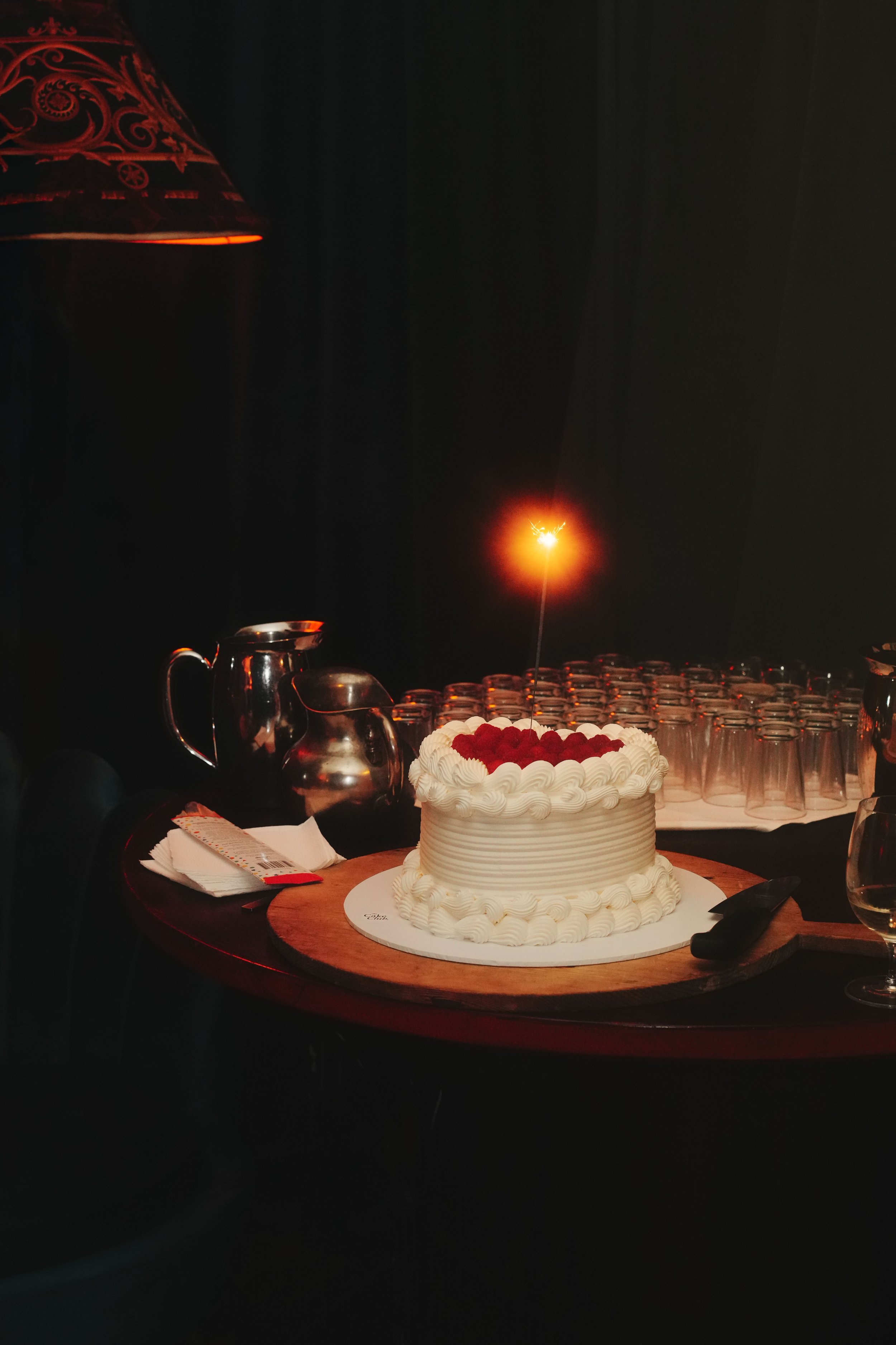 A white frosted cake decorated with red berries and piped icing on a wooden board, with a sparking firework candle on top, placed on a table with glassware and utensils in the background.