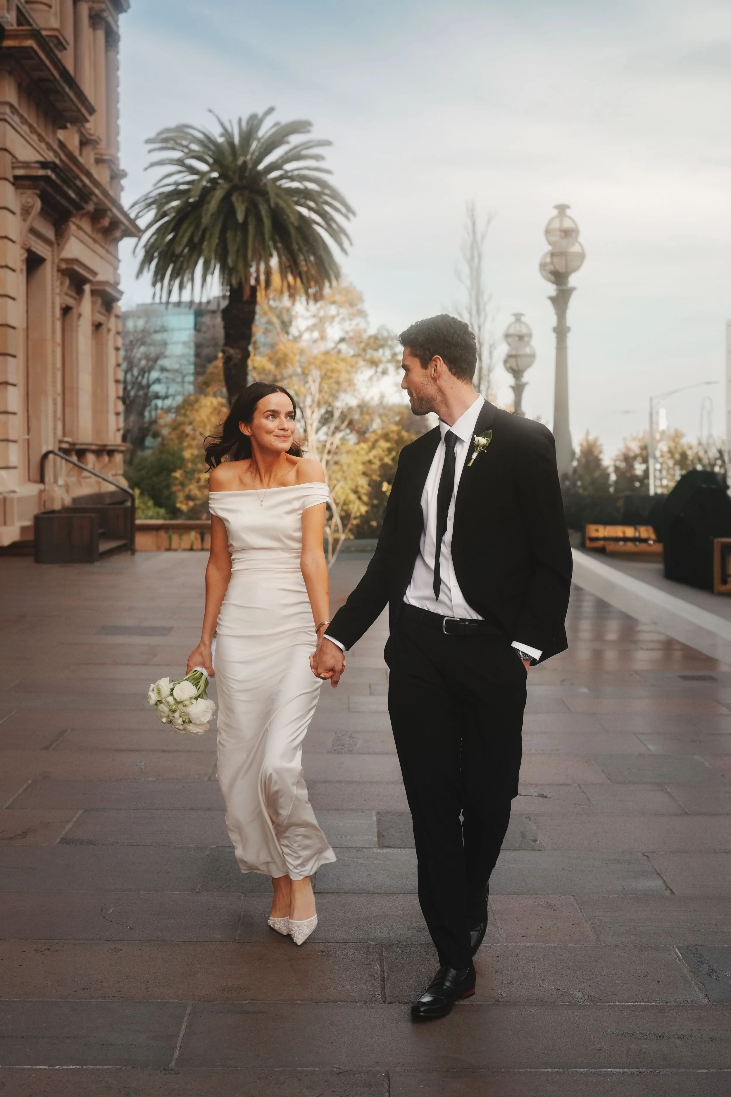 A bride and groom walking hand in hand outdoors during their wedding. The bride is wearing a white off-shoulder wedding dress and holding a bouquet of white roses. The groom is dressed in a black suit with a white shirt and black tie, with a boutonni
