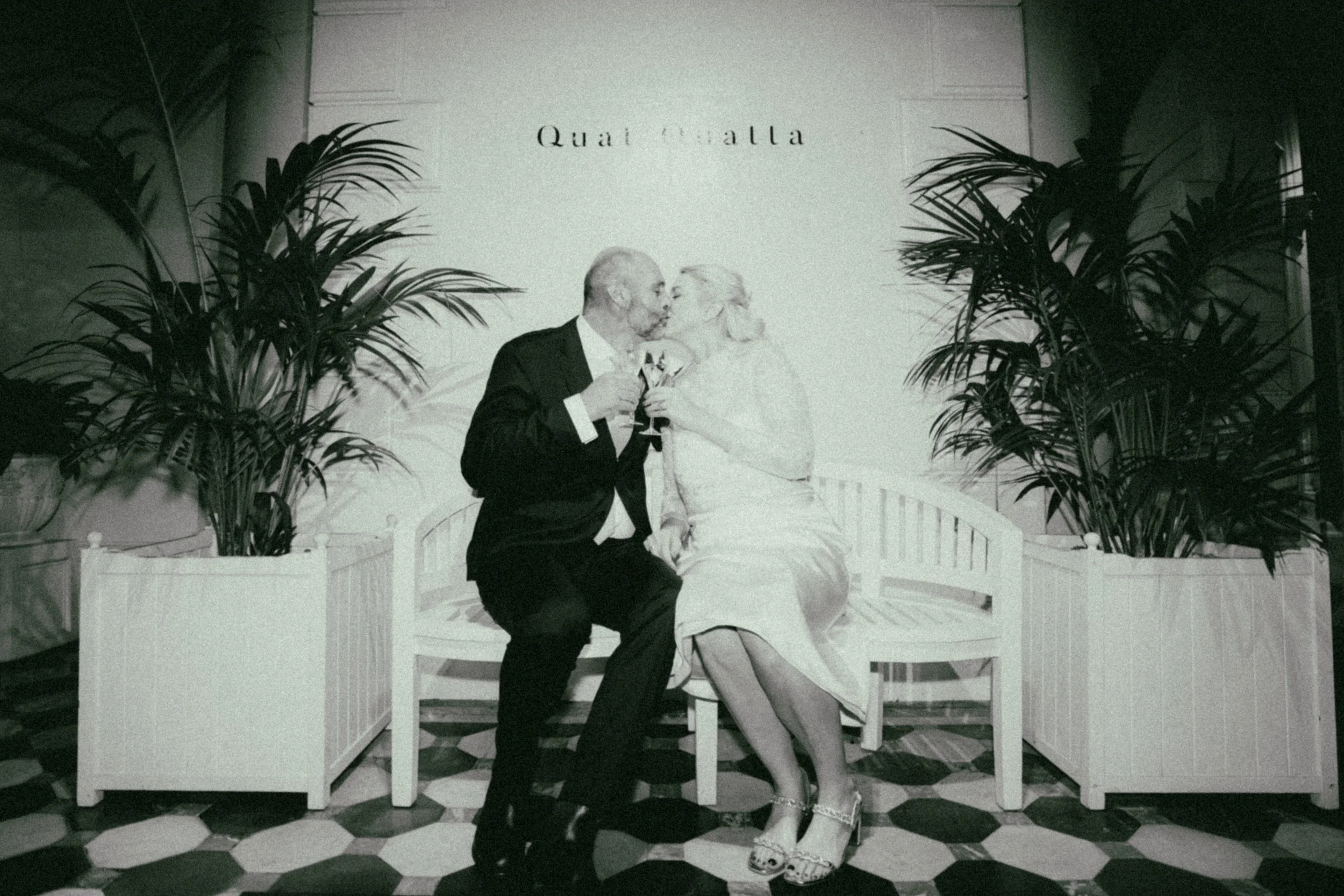 An elderly man and woman are sitting on a white bench, sharing a kiss and holding glasses of wine. The man is dressed in a suit and tie, while the woman is wearing a dress and heels. There are large plants on either side of the bench, and a sign that