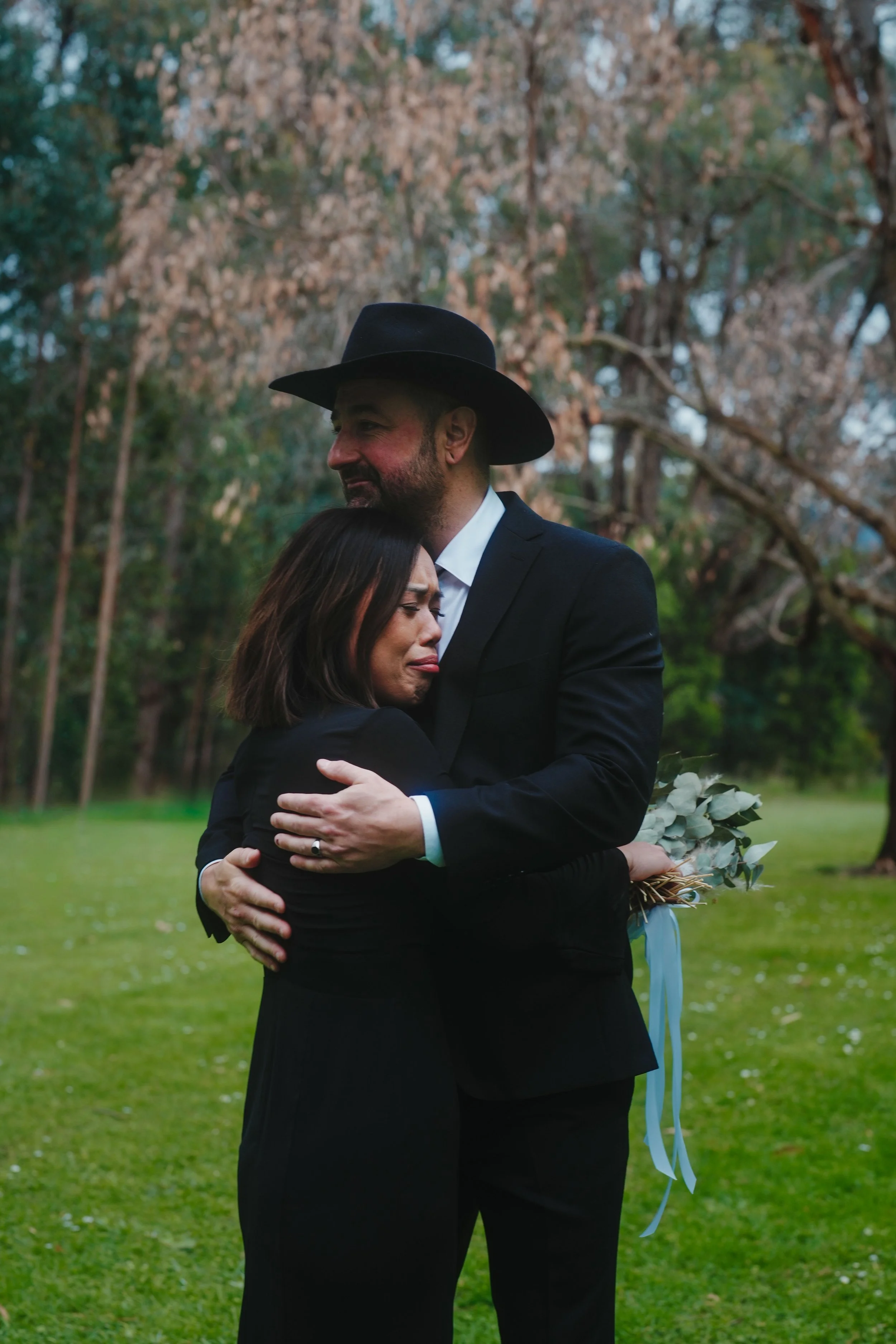 A man in a black suit and wide-brimmed hat hugs a woman in a black dress, holding a bouquet of eucalyptus leaves tied with a blue ribbon, outdoors with trees and grass in the background.