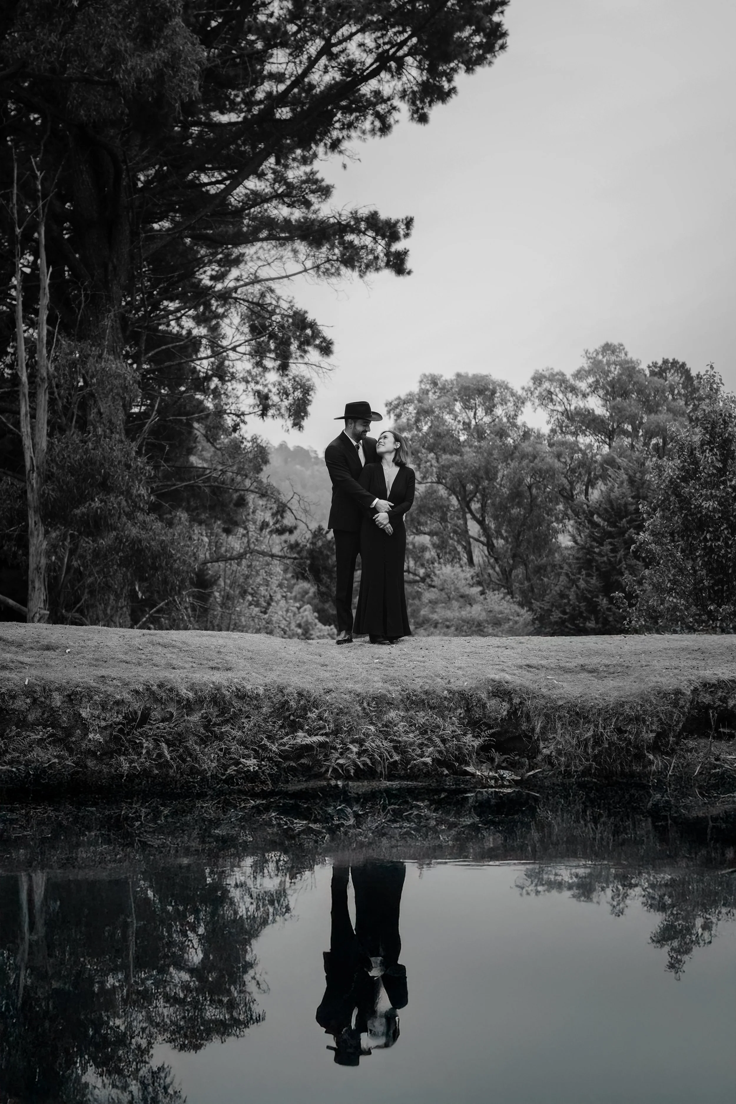 Black and white photograph of a couple standing outdoors near a calm body of water, with trees and cloudy sky in the background. The man is wearing a hat and suit, and the woman is dressed in a long dress. They are gazing at each other, and their ref