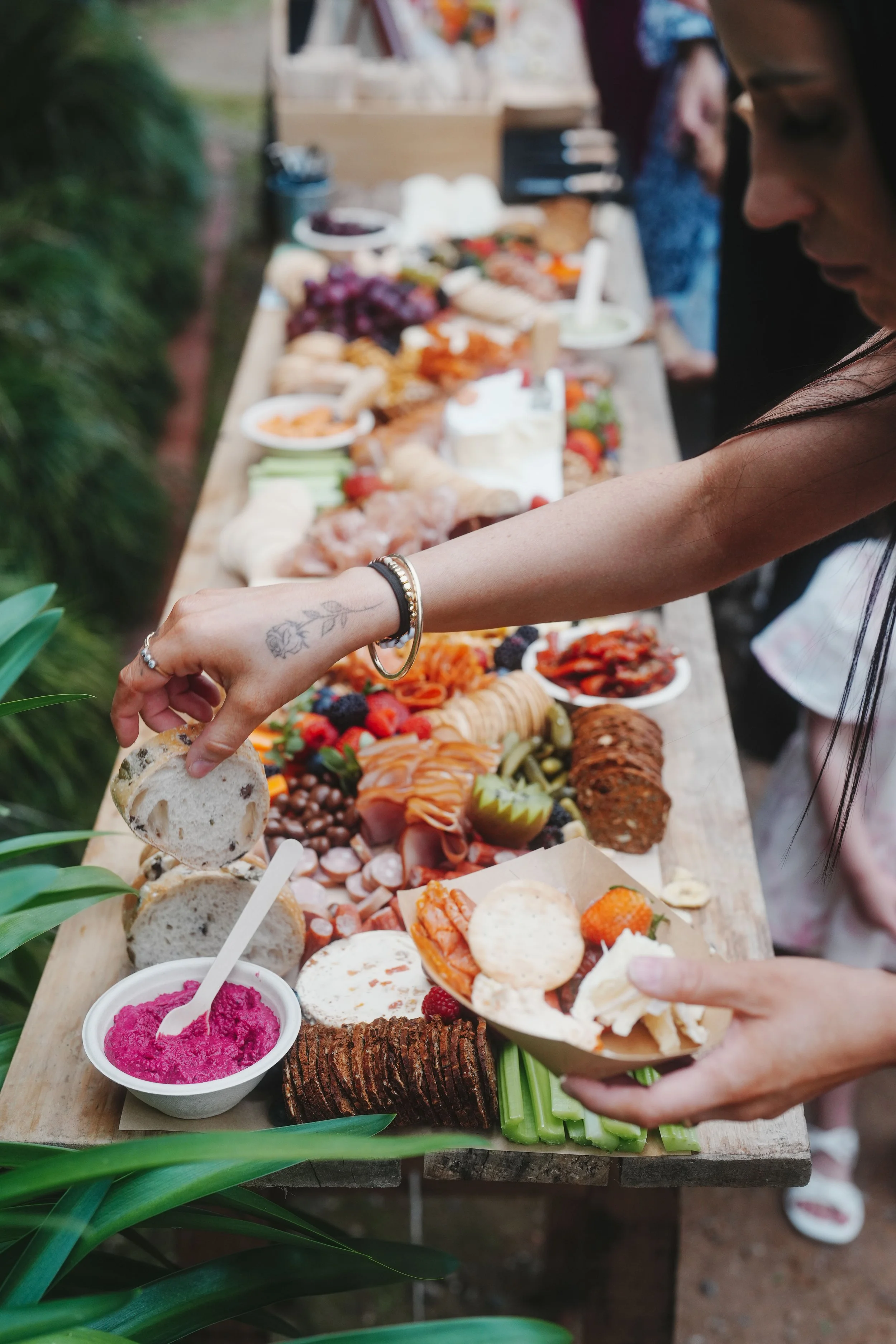 A woman is reaching for a piece of bread at an outdoor buffet table filled with various foods, including bread, desserts, fruits, and spreads, with green plants in the foreground.