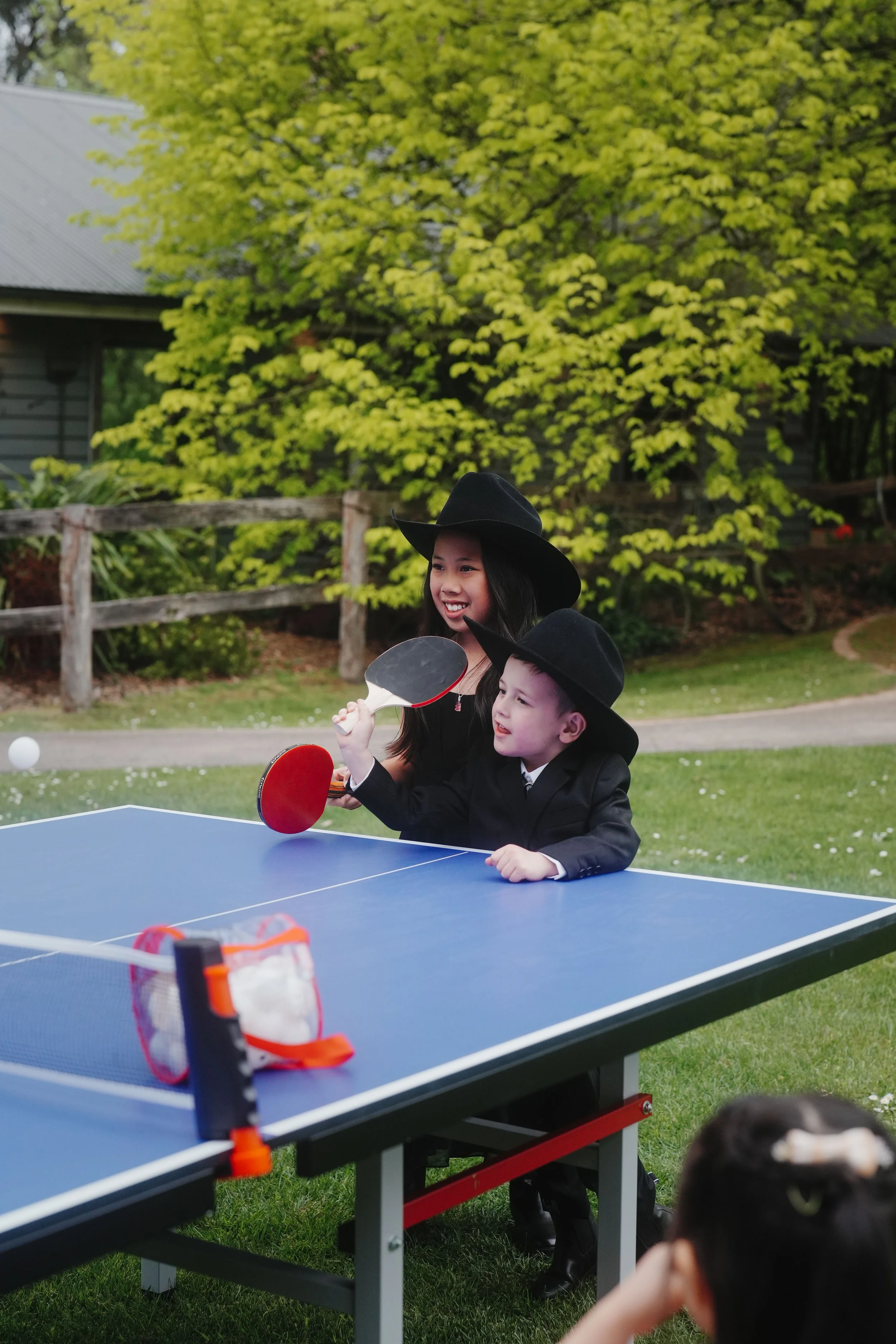 Two children playing table tennis outdoors on a sunny day with trees and a house in the background. The girl is smiling and holding a paddle, the boy is also playing with a paddle. Both are wearing black hats and dark clothing.