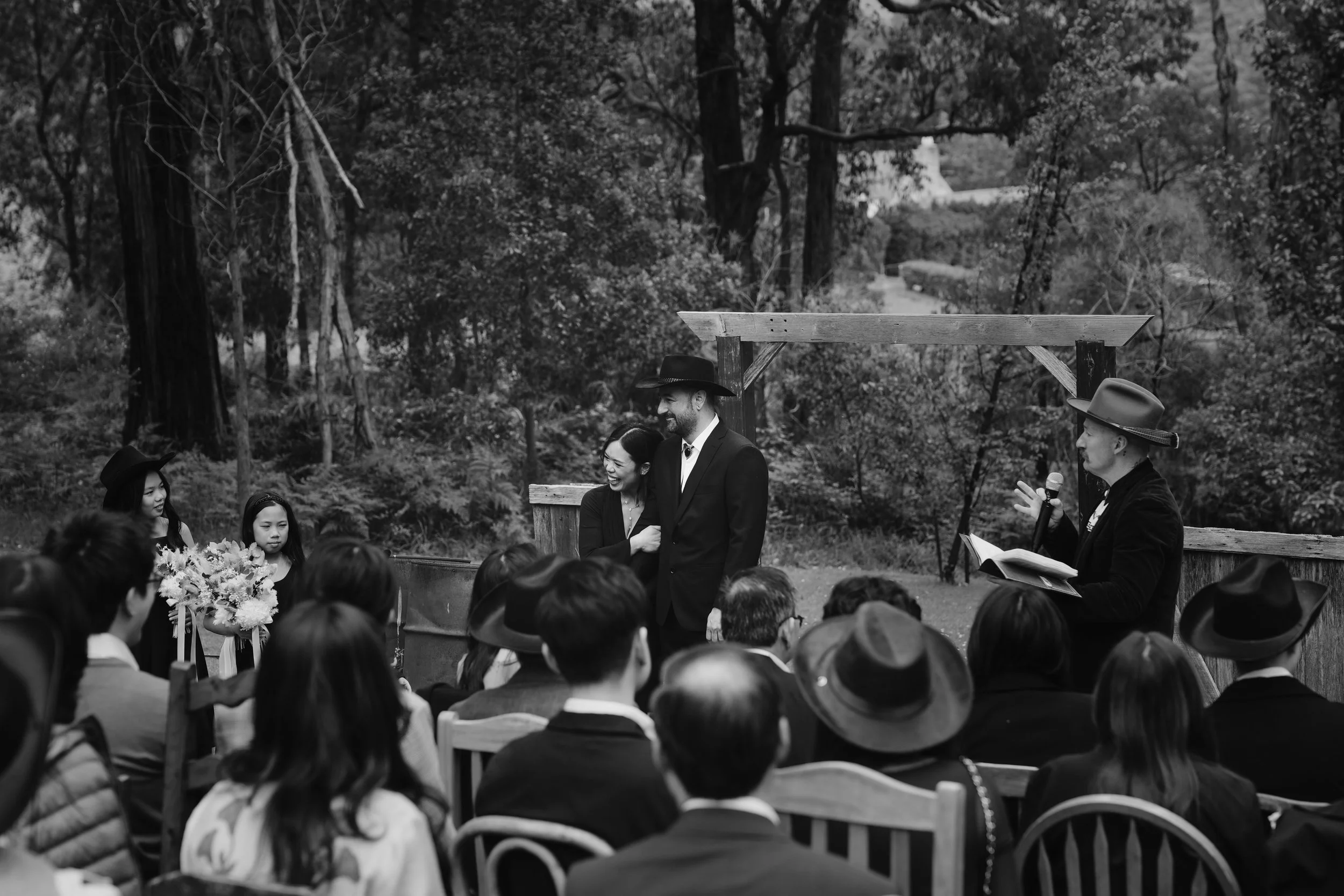 A black and white photo of an outdoor ceremony in a natural setting with trees. A man and woman are standing in front of an audience, smiling at each other. The man is wearing a cowboy hat, and the woman is holding a bouquet of flowers. Someone is re
