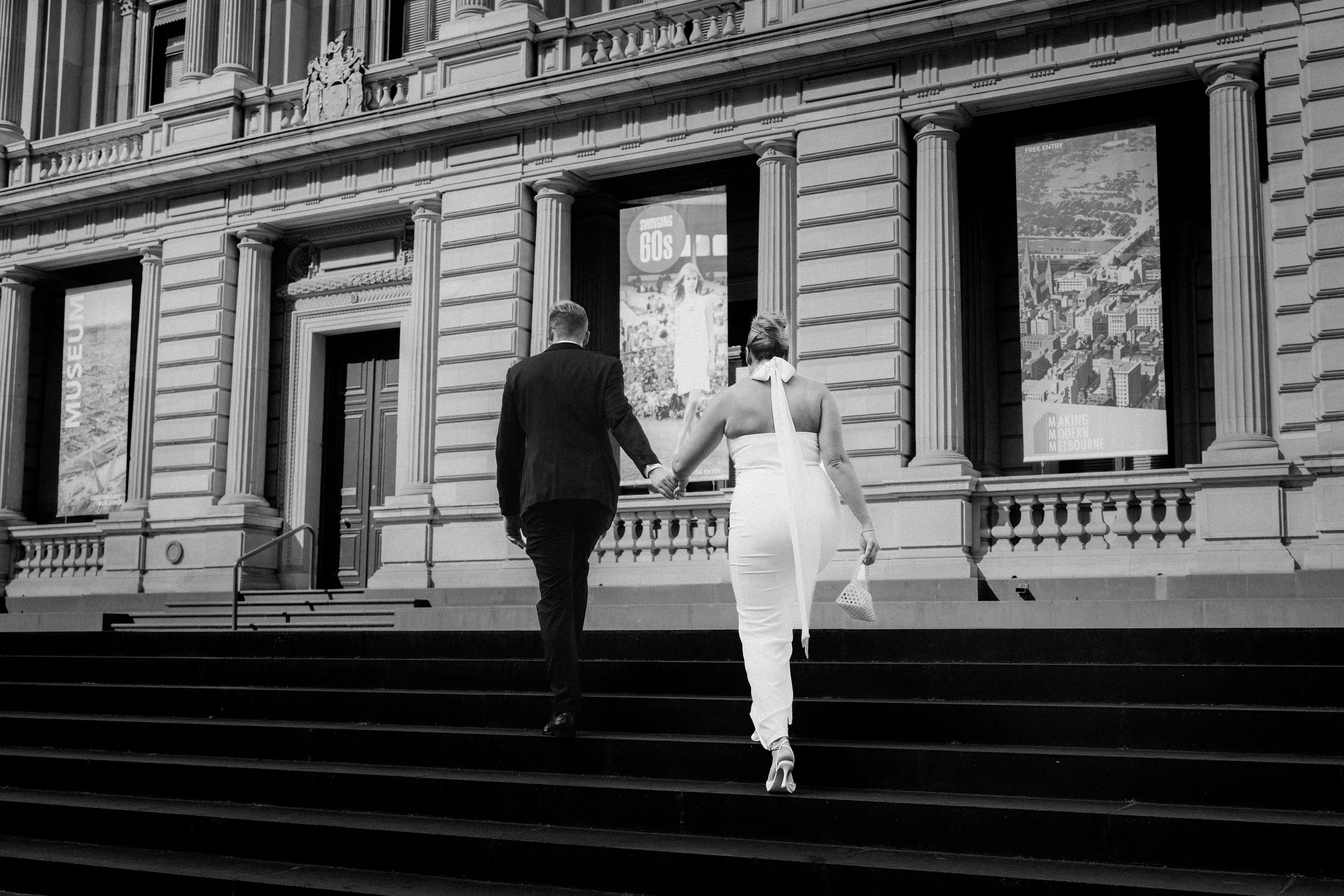 A black and white photo of a man and woman in formal attire walking up the steps of a historic building with ornate architectural details and large posters in the windows.