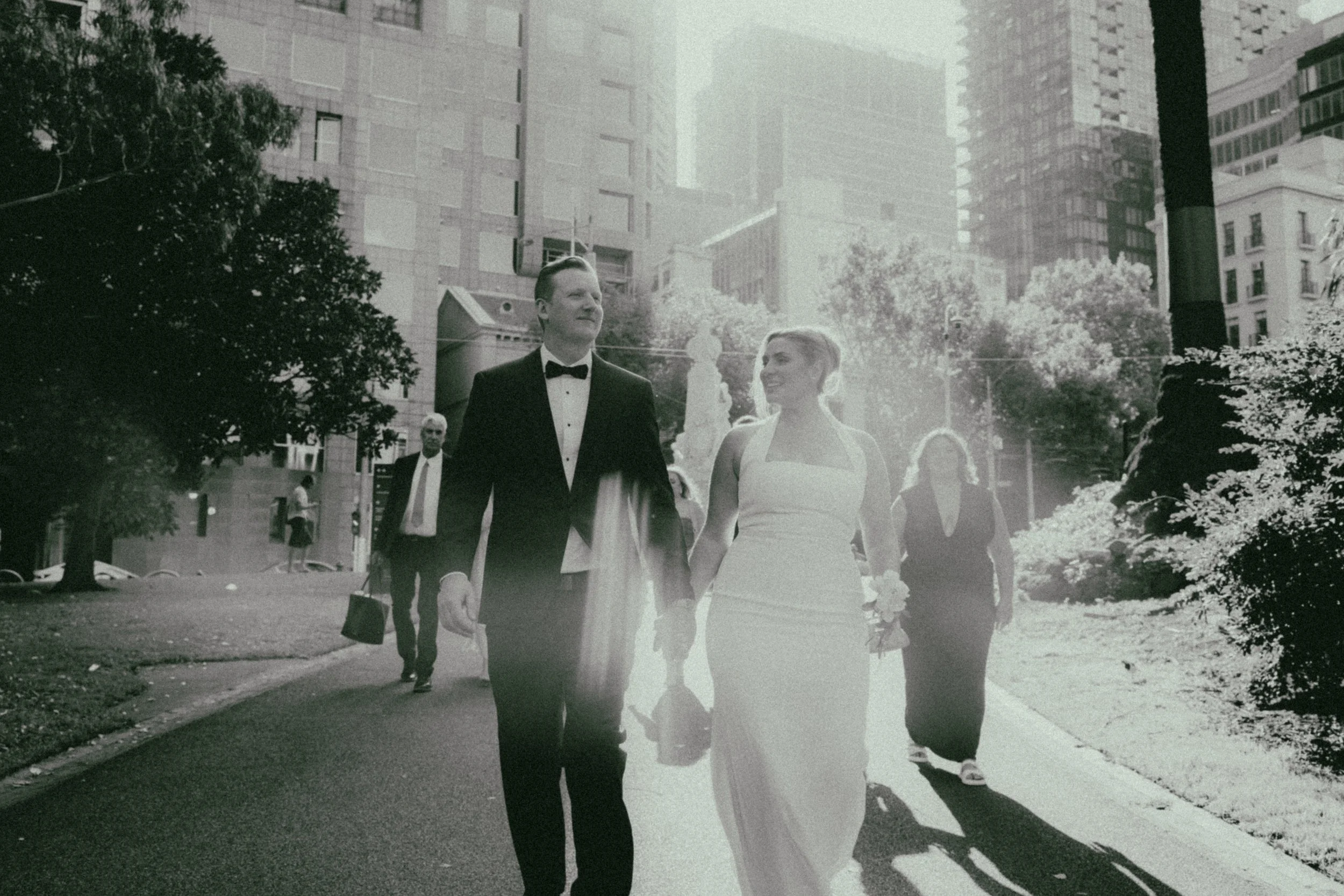 A black-and-white photo of a man in a tuxedo and a woman in a wedding dress walking together in city park, surrounded by tall buildings and trees, with other people in the background.