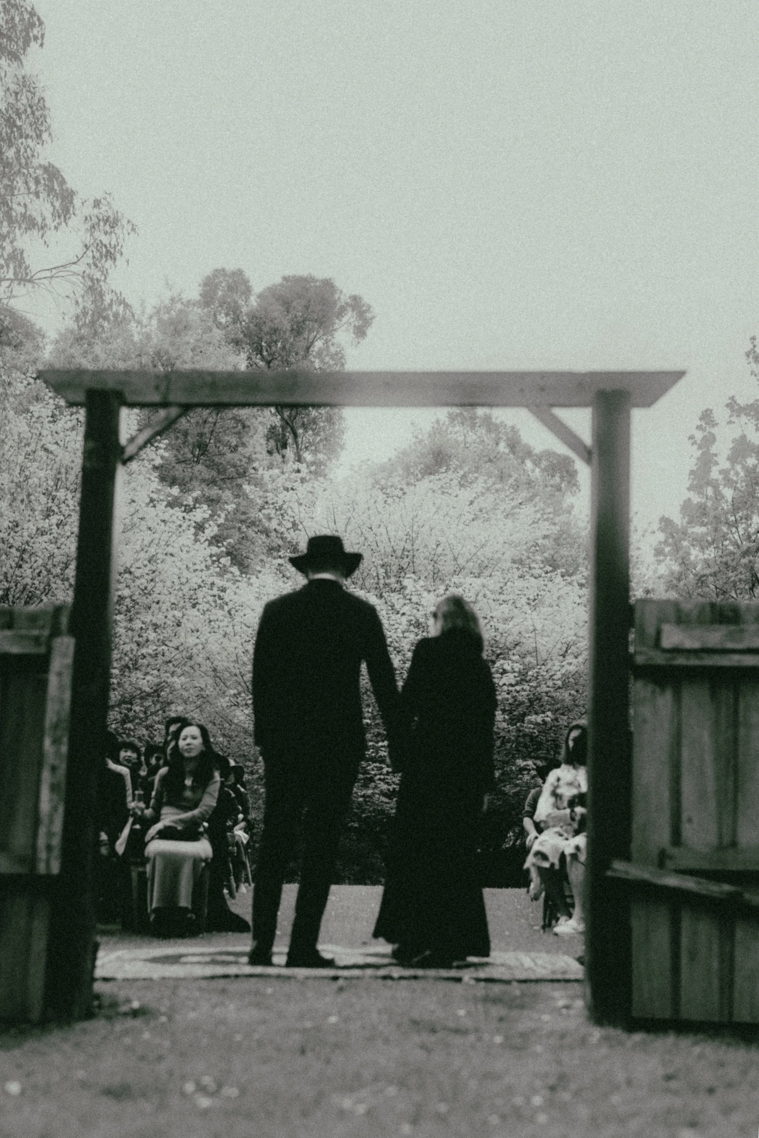 A black and white photograph of a group of people, including children and adults, seated and standing in an outdoor setting surrounded by trees. Two individuals, one man and one woman, are walking away from the camera through a wooden entrance, holdi