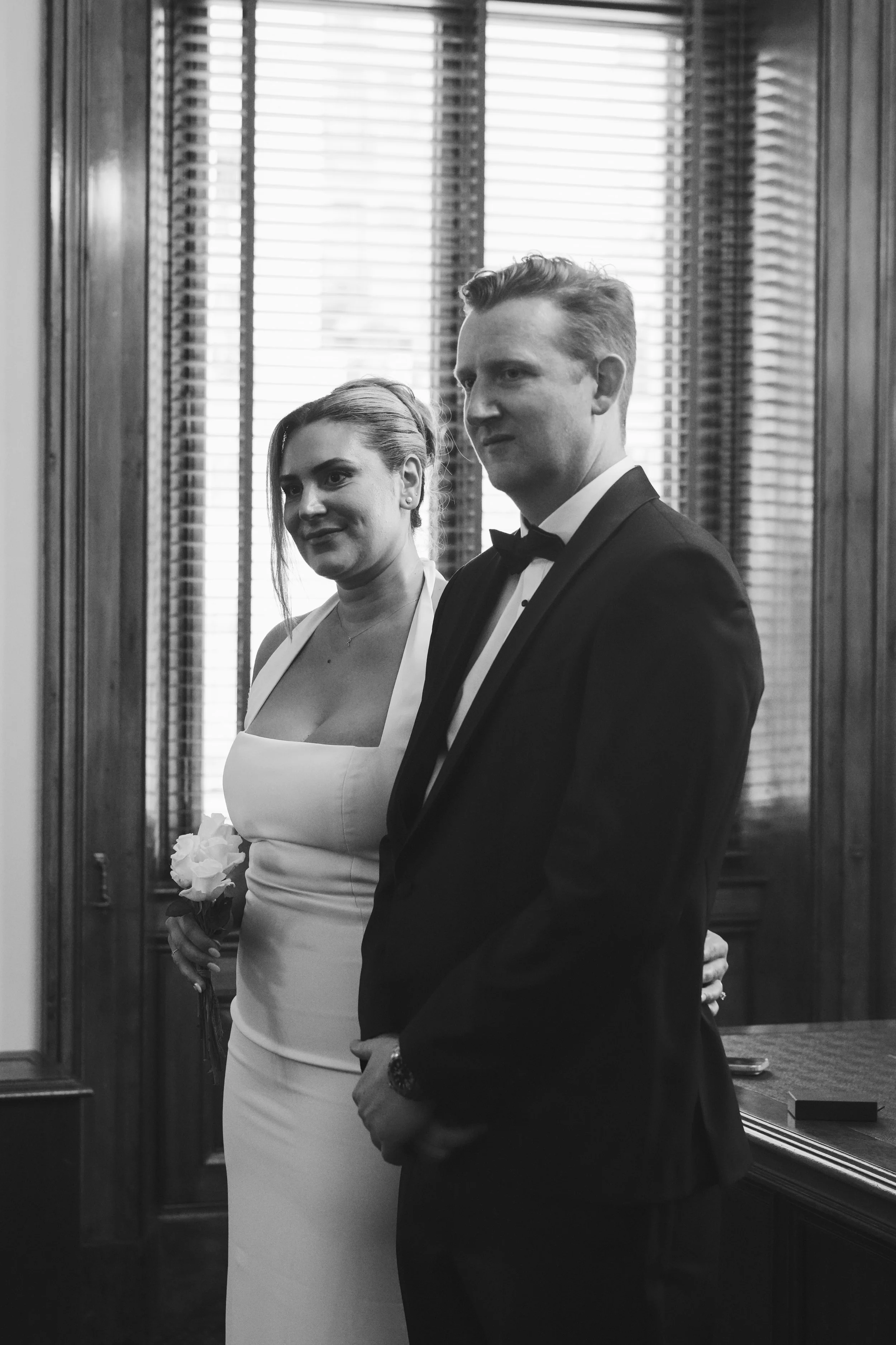 A black and white photo of a woman in a white dress holding a flower, standing close to a man in a tuxedo with a bow tie. They are indoors near a window with wooden shutters, both looking to the side.