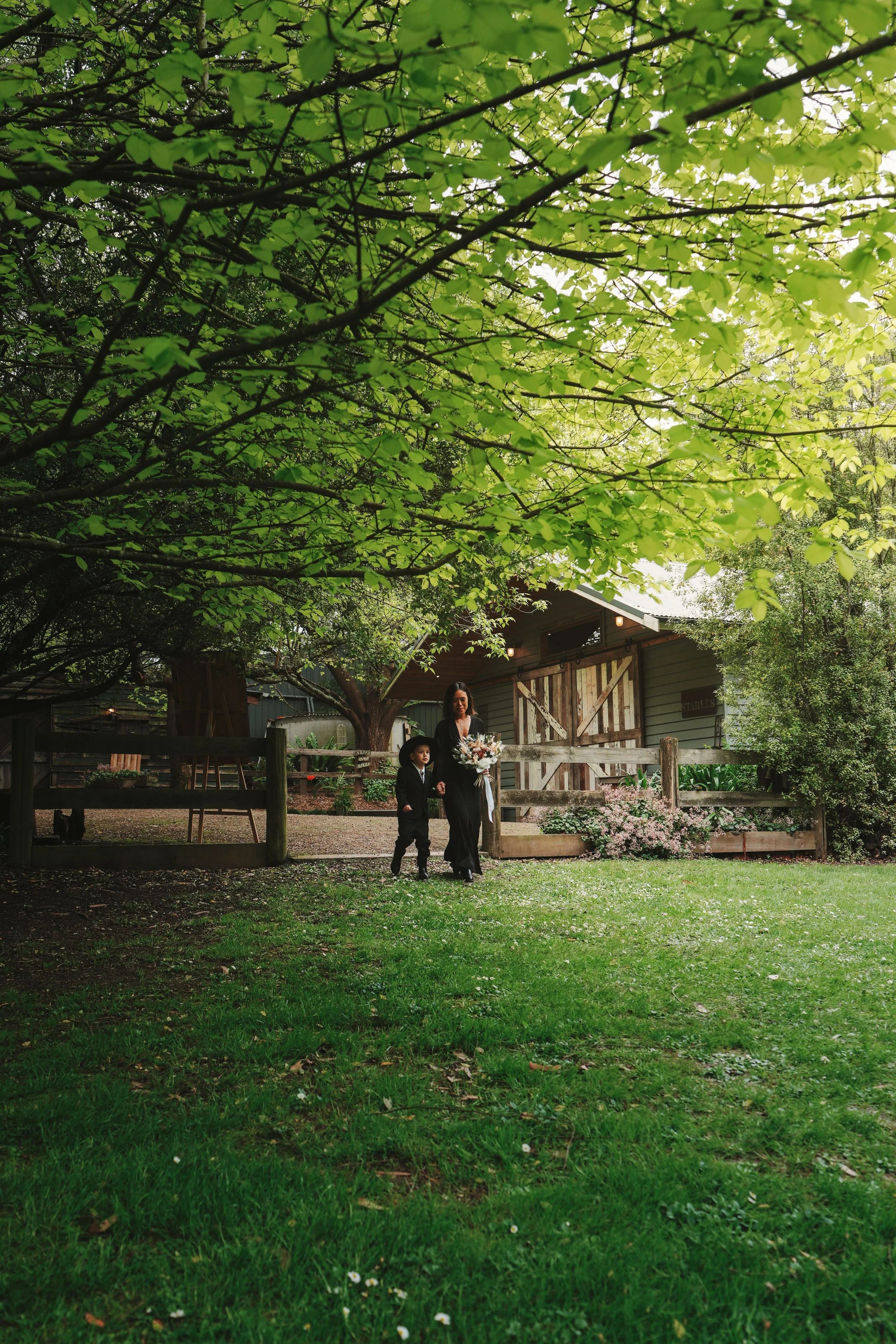 A woman and a young boy walk together in a garden filled with trees, flowers, and a wooden fence. The woman holds a bouquet of flowers and the boy wears a suit. In the background, there is a rustic building with a barn door.