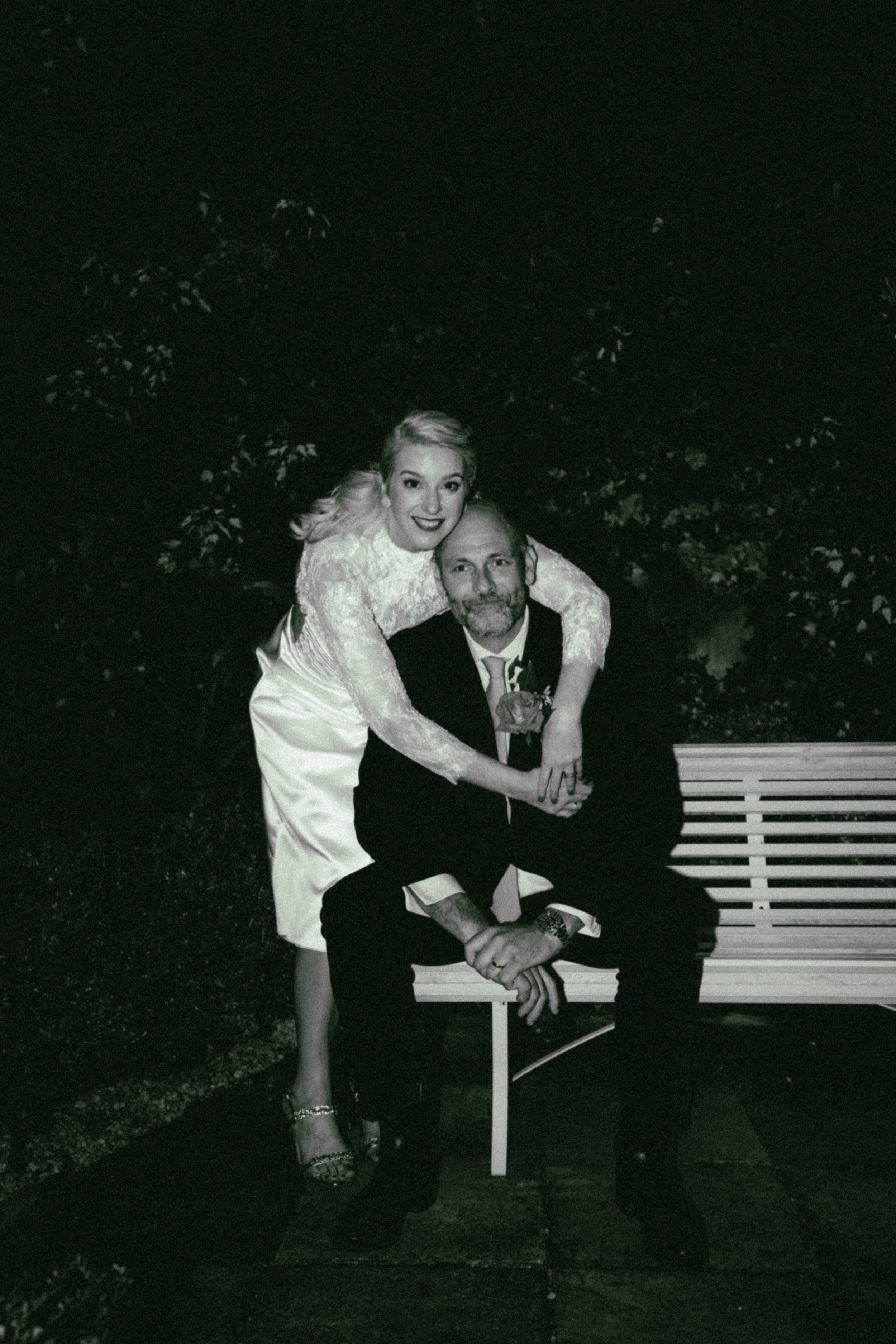 A black and white photo of a woman and man sitting on a park bench at night. The woman is standing behind the man, embracing him with her arms around his shoulders, and smiling at the camera. The woman is wearing a long dress and high heels, while th