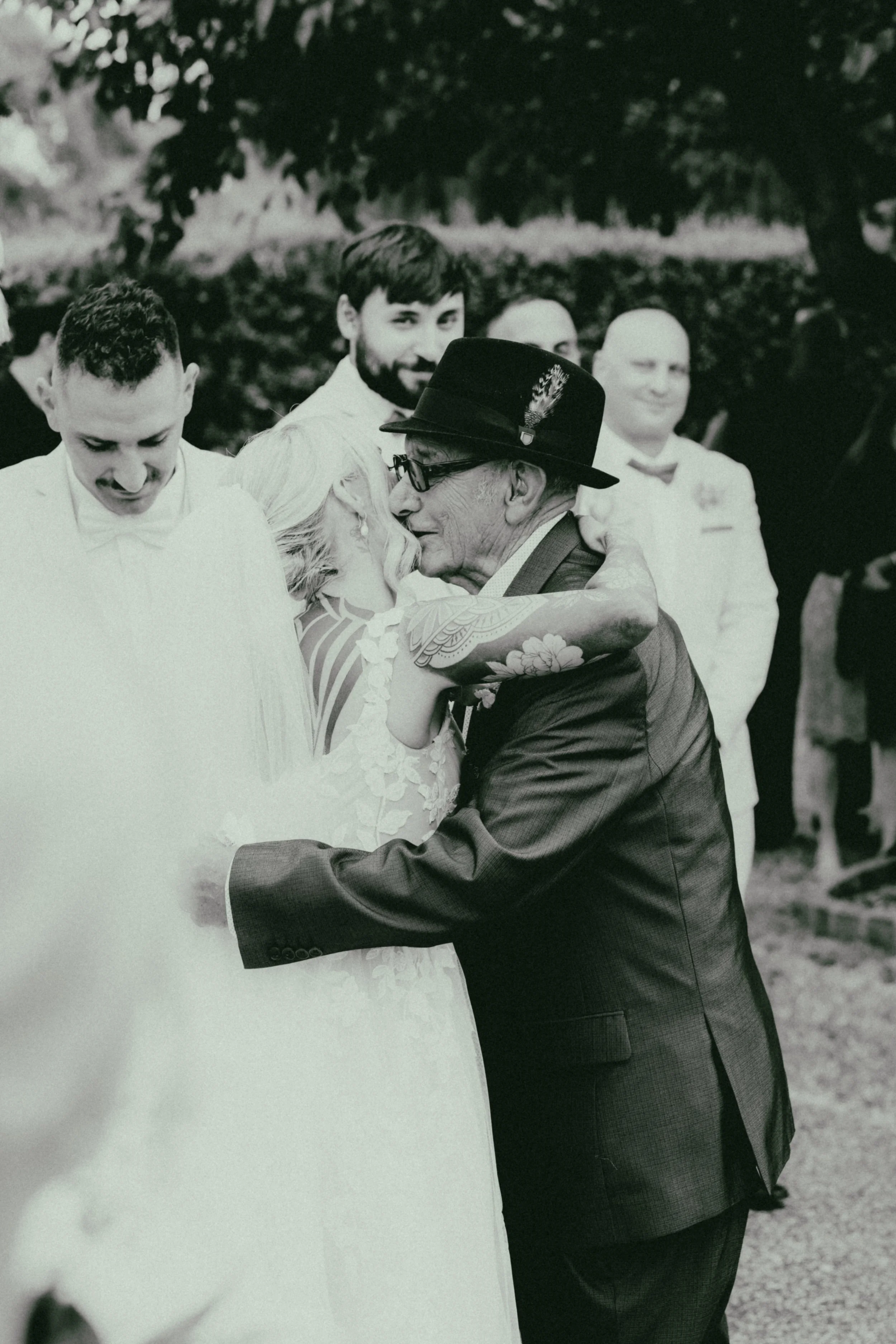 An elderly man in a suit and hat embracing a woman in a wedding dress, with a group of people in the background at an outdoor event.