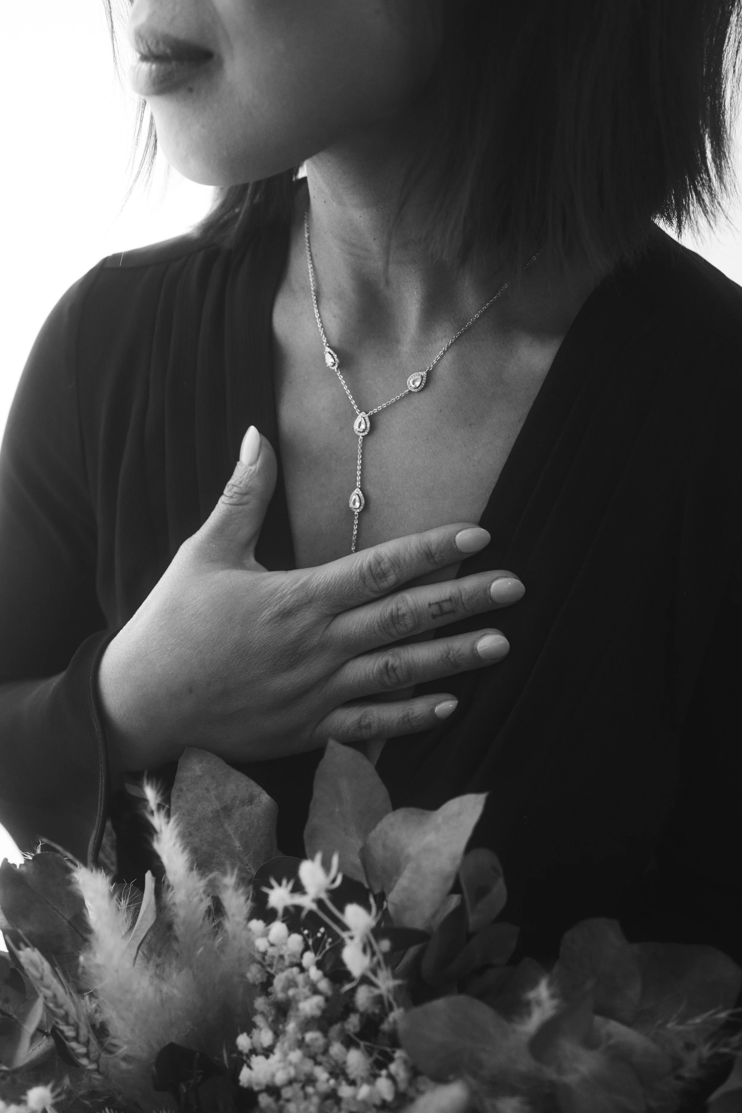 A close-up black and white photo of a woman with shoulder-length dark hair wearing a necklace with teardrop-shaped pendants. She is dressed in a dark top and places one hand on her chest. The background shows a bouquet of flowers with various leaves 
