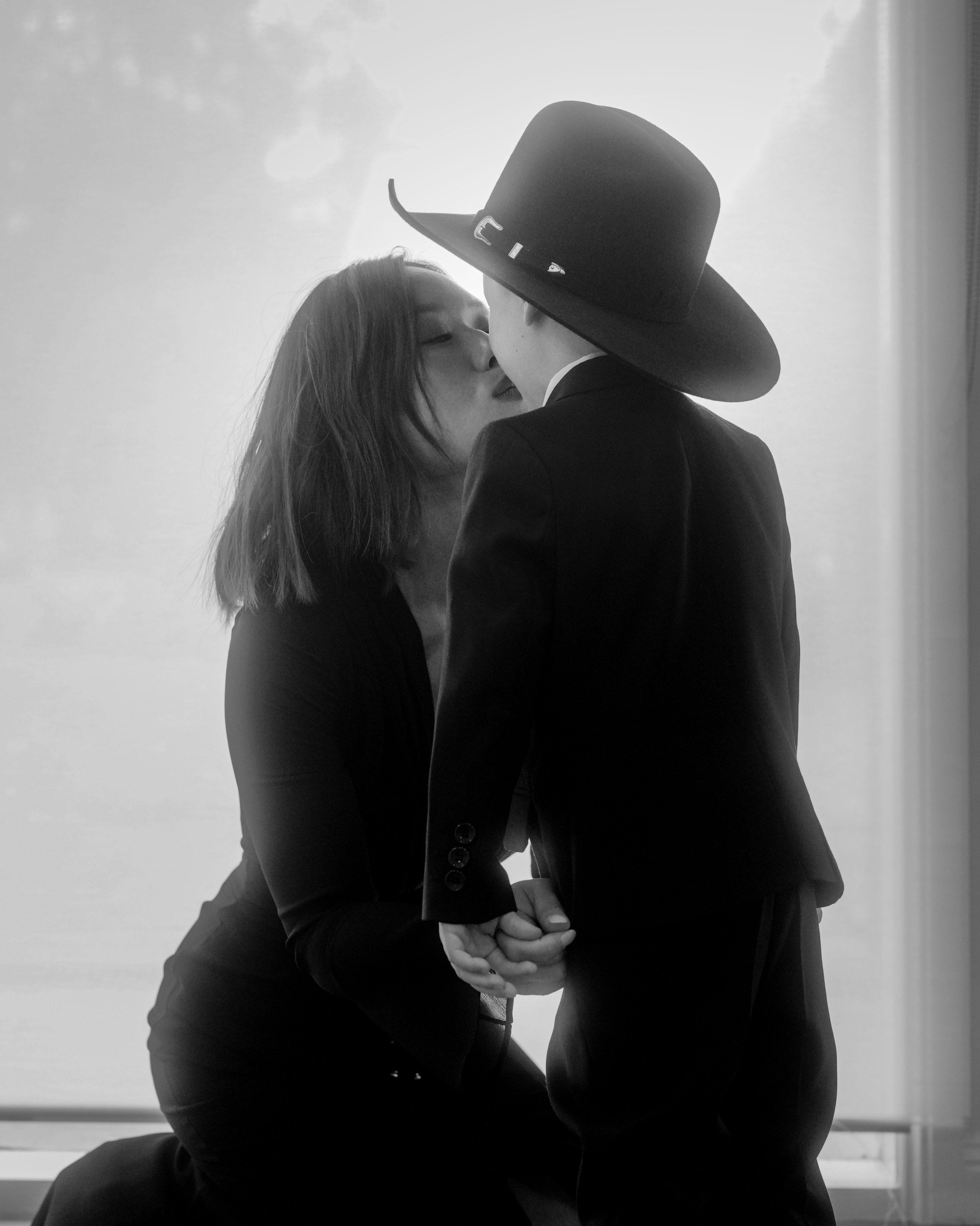 A black and white photo of a man and woman in formal clothing kissing, with the woman sitting and the man standing under a wide-brimmed hat, indoors near a window.