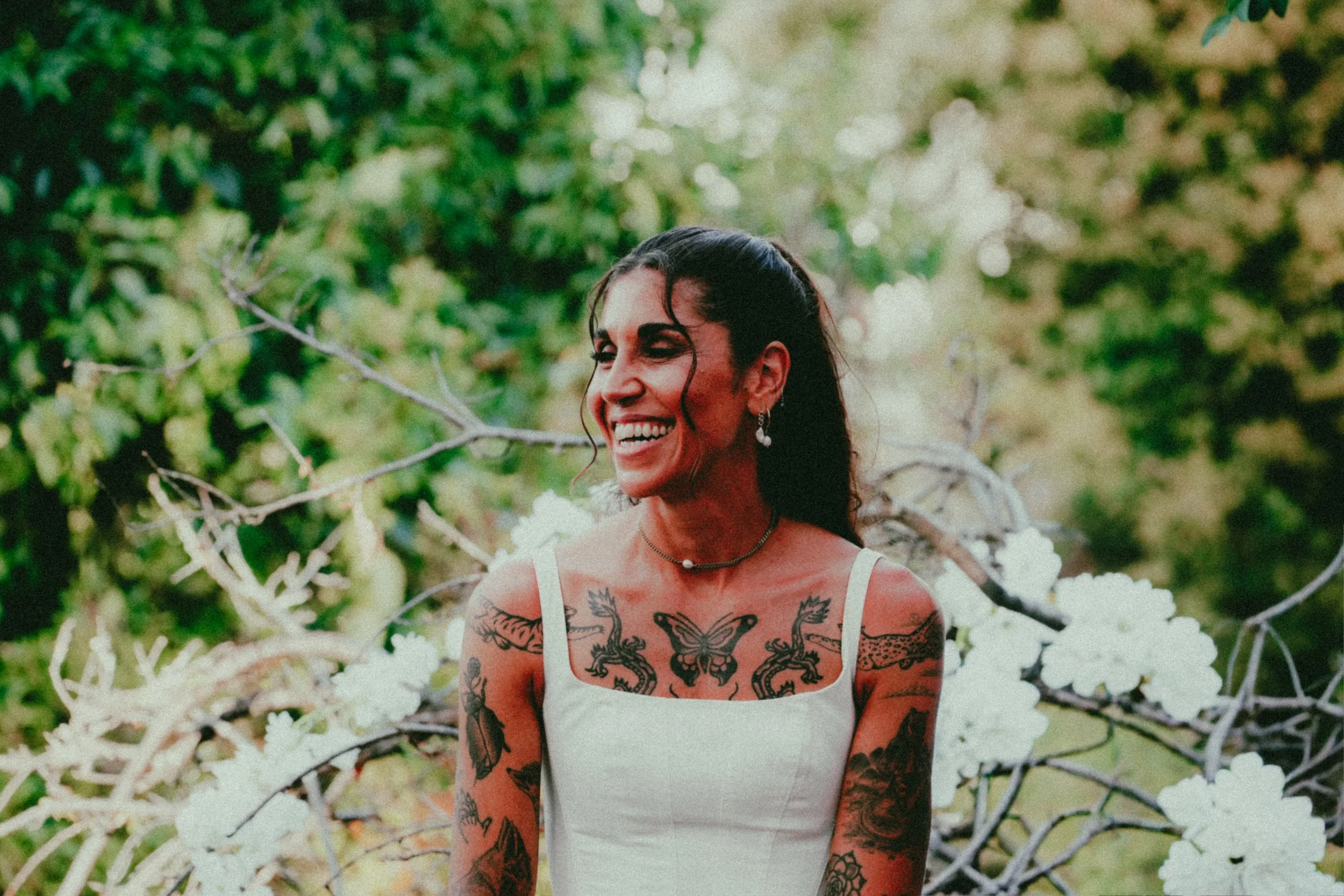 A smiling woman with dark hair and tattoos on her arms and chest, wearing a white tank top, standing amid green foliage and white flowers.