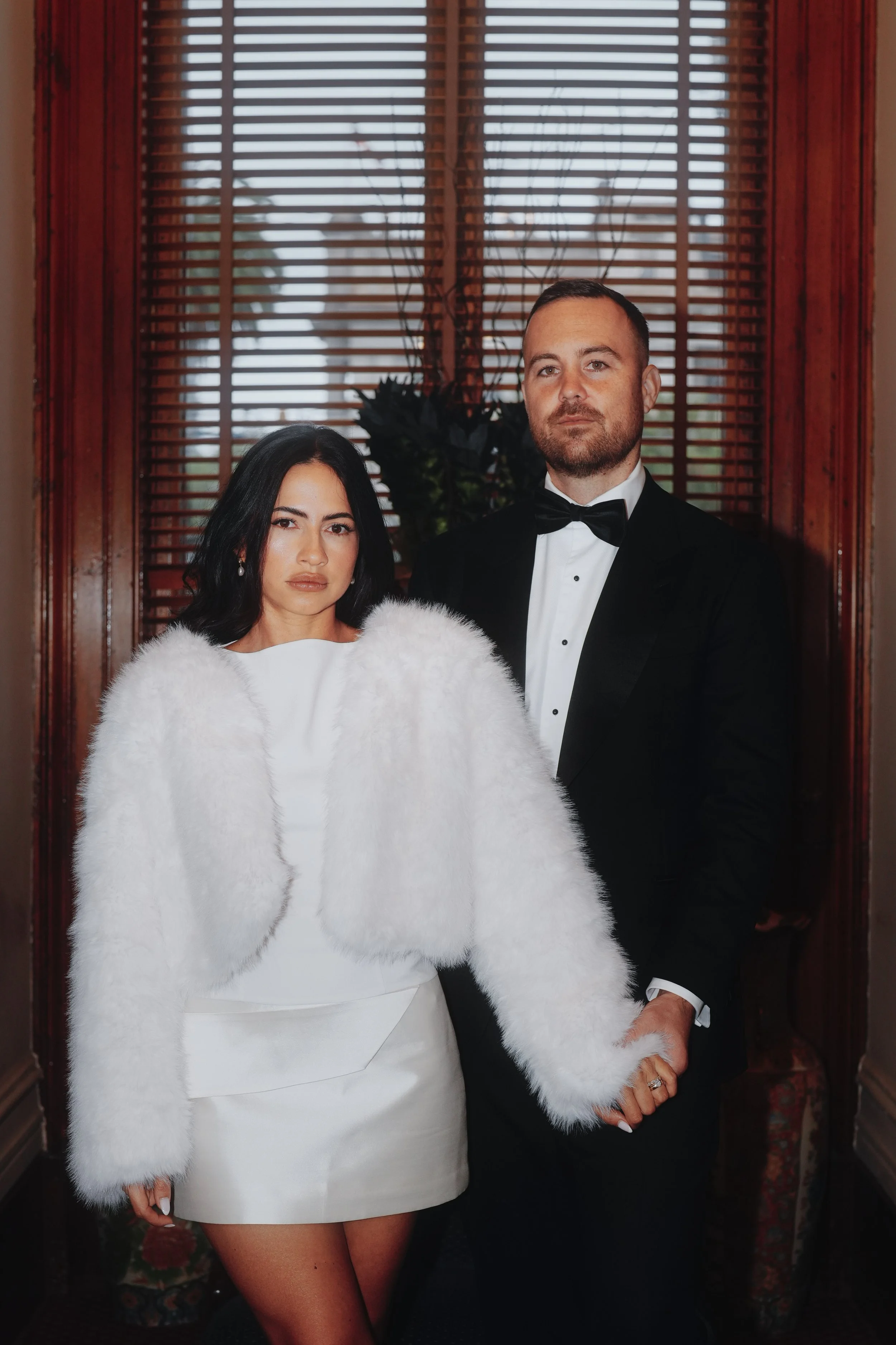 A man and woman dressed formally, holding hands, standing in front of window blinds and a potted plant. The woman is wearing a white dress with a white fur stole, and the man is in a black tuxedo with a bow tie.