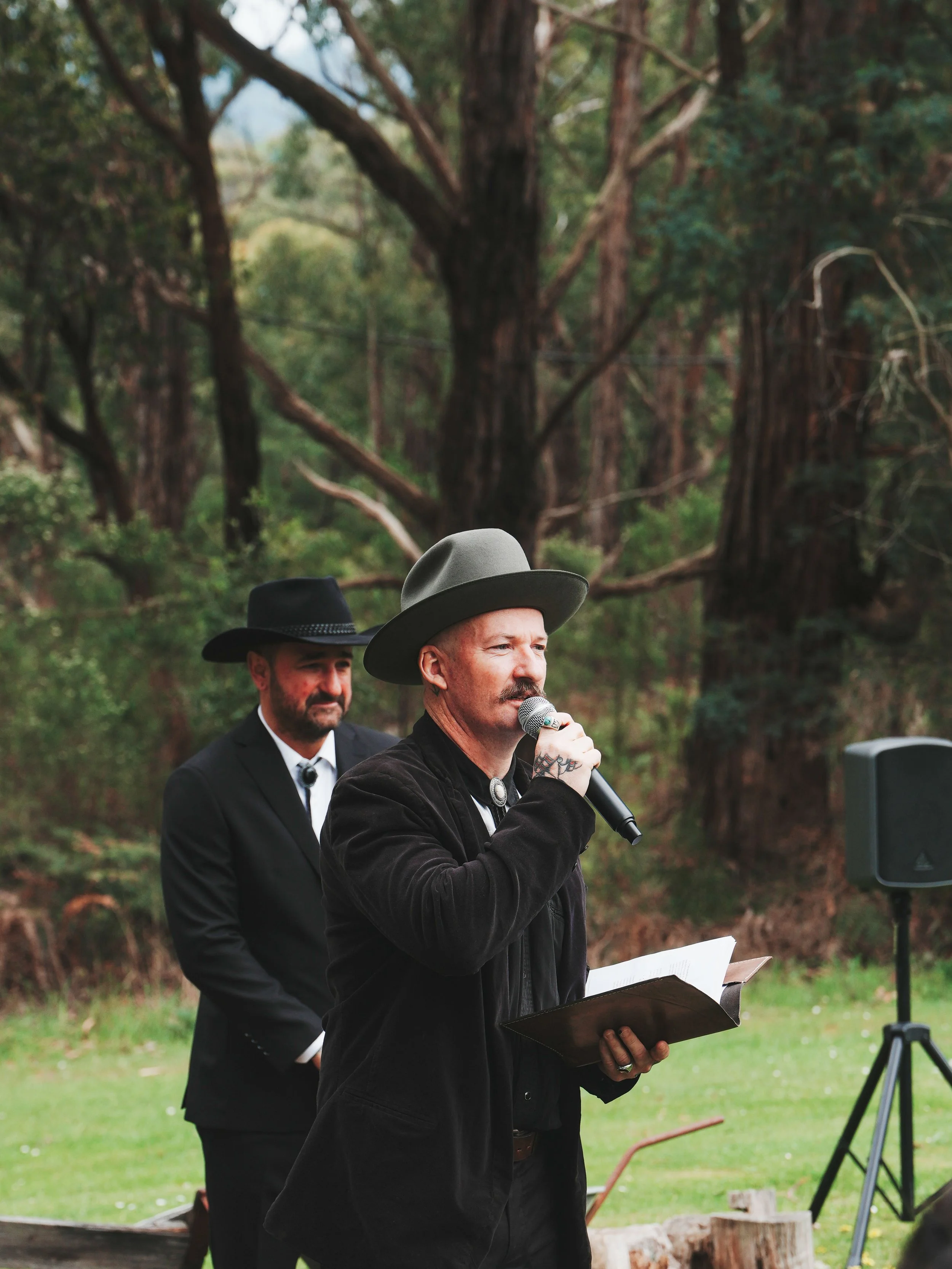 A man with a mustache and tattoo on his neck wearing a gray hat, speaking into a microphone during an outdoor event in a wooded area, with two men dressed in black suits and hats in the background.