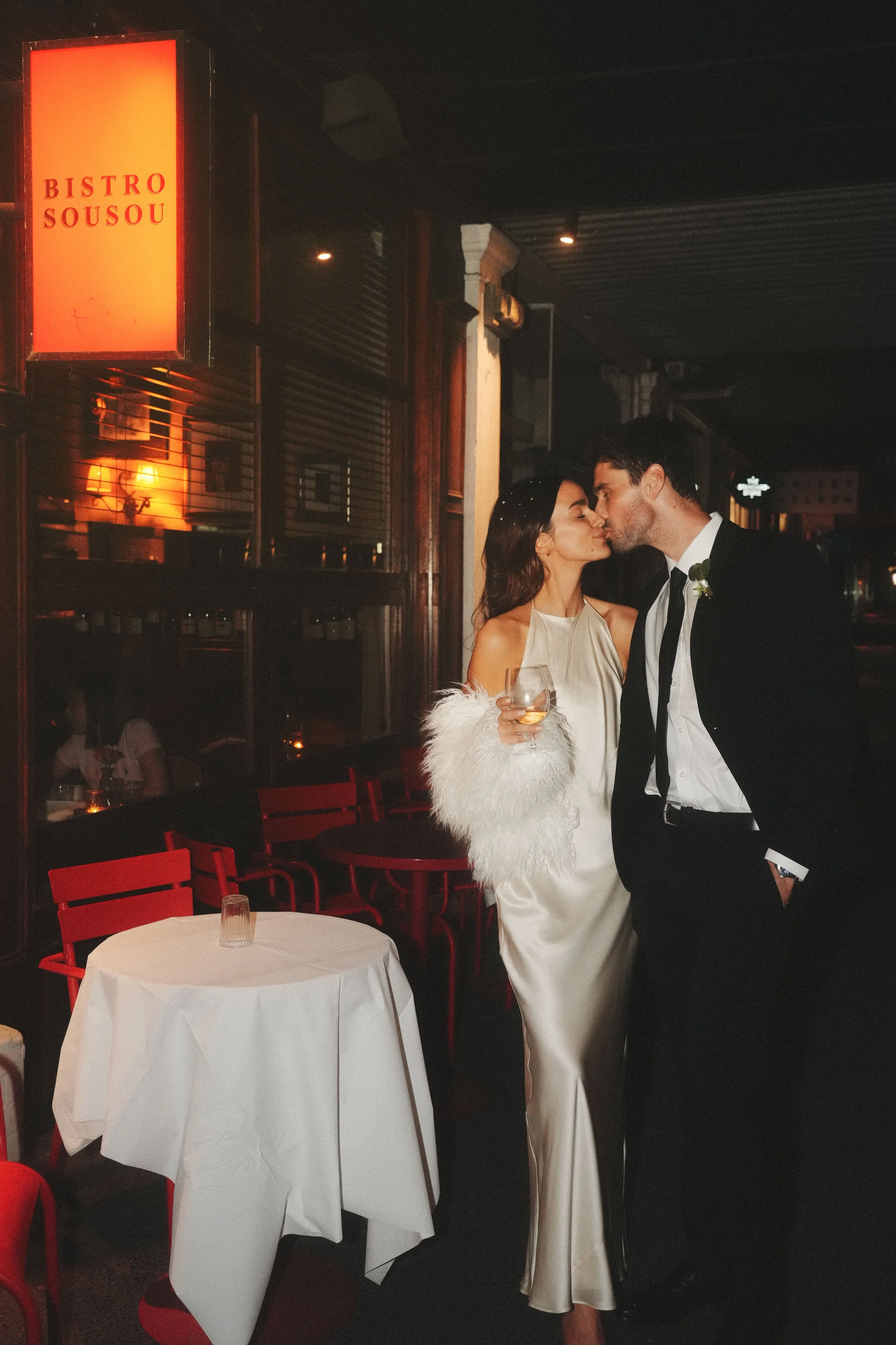 A couple dressed in formal attire, the woman in a white gown and the man in a black tuxedo, sharing a kiss at a restaurant. The woman holds a glass of white wine. The restaurant has red chairs, white tablecloths, and dark lighting with warm glow from