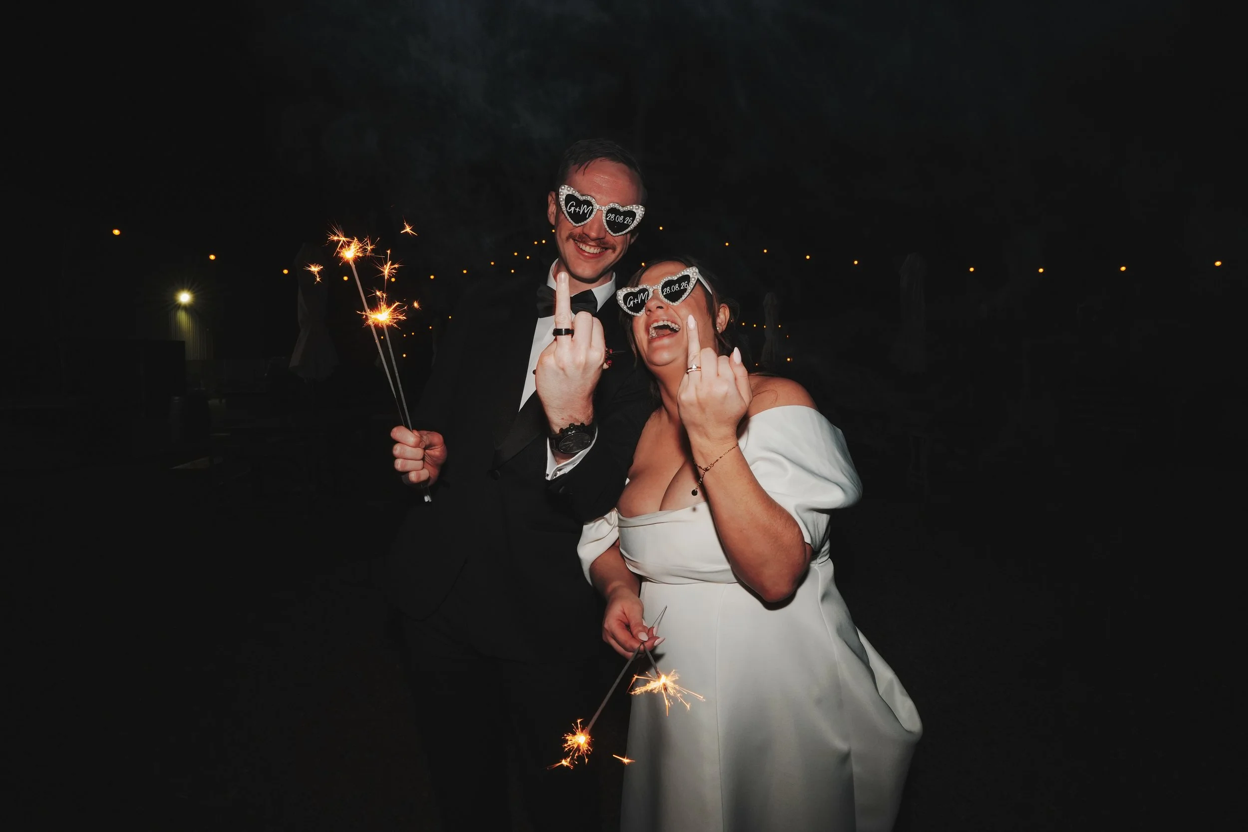 A newlywed couple celebrating at night, holding sparklers and wearing sunglasses with wedding details, smiling and making gestures, with a dark sky and string lights in the background.