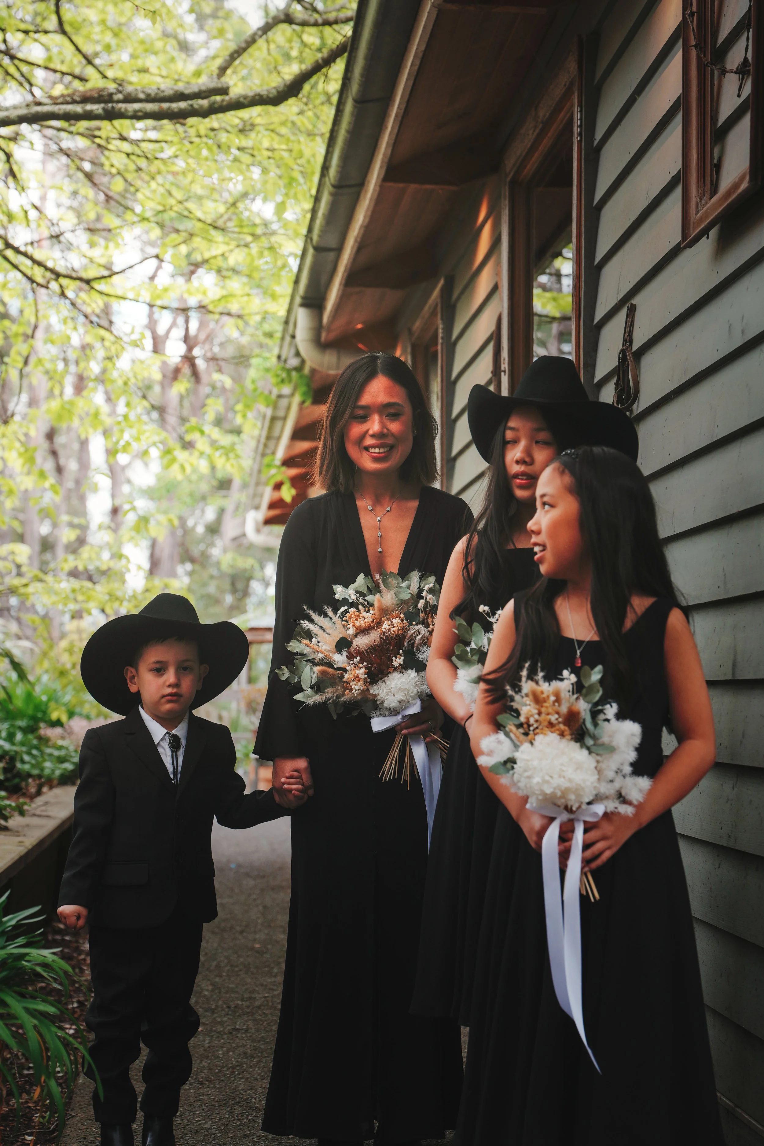 Group of people dressed in formal black attire, standing outdoors, holding bouquets of flowers, on a narrow path with trees and a wooden building in the background.