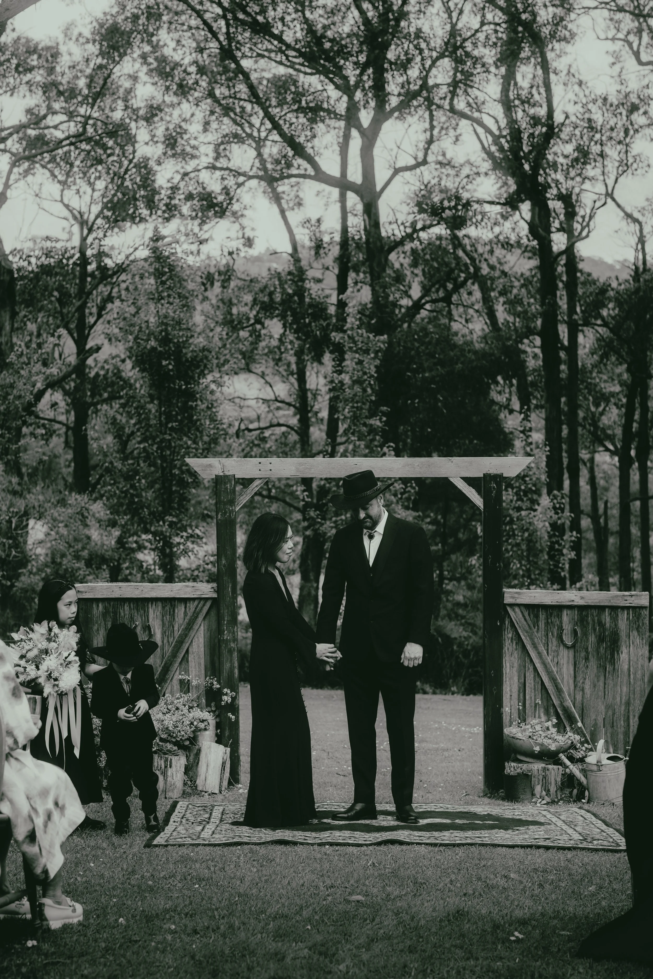 A black and white photo of a wedding ceremony outdoors. A couple is holding hands and looking at each other, with an officiant standing in front of them. Several children are seated nearby as witnesses, and a floral arrangement with ribbons is visibl