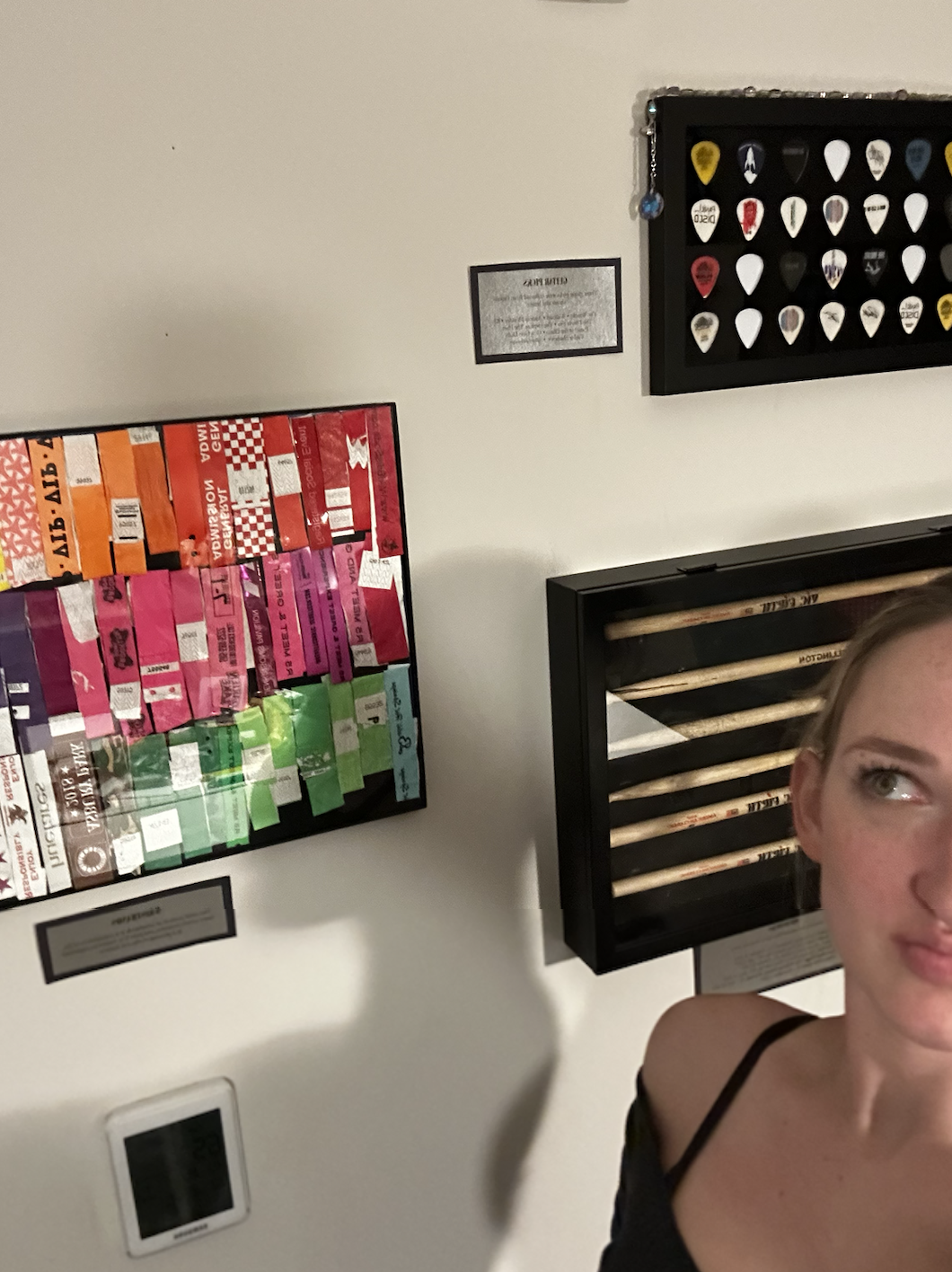Partial view of a woman with blonde hair and a black strap top, standing near a wall with framed items, including a display of guitar picks and a colorful collage of event or ticket stubs.