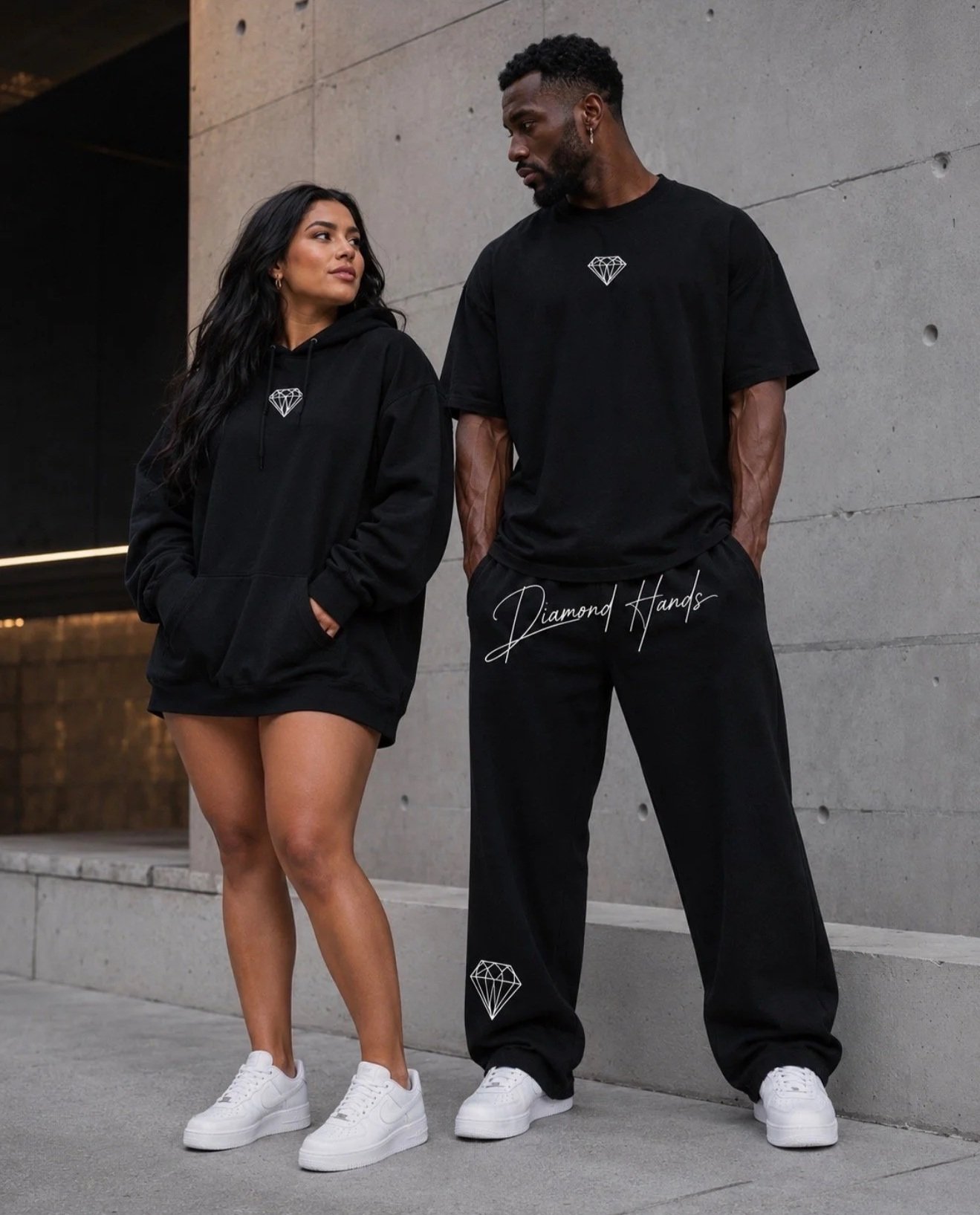 A woman and a man dressed in black casual clothing, standing against a concrete wall. The woman wears a black hoodie with a diamond logo and white sneakers, while the man wears a black t-shirt with a diamond logo, black pants, and white sneakers. The text "Diamond Hands" is written on the man's pants.